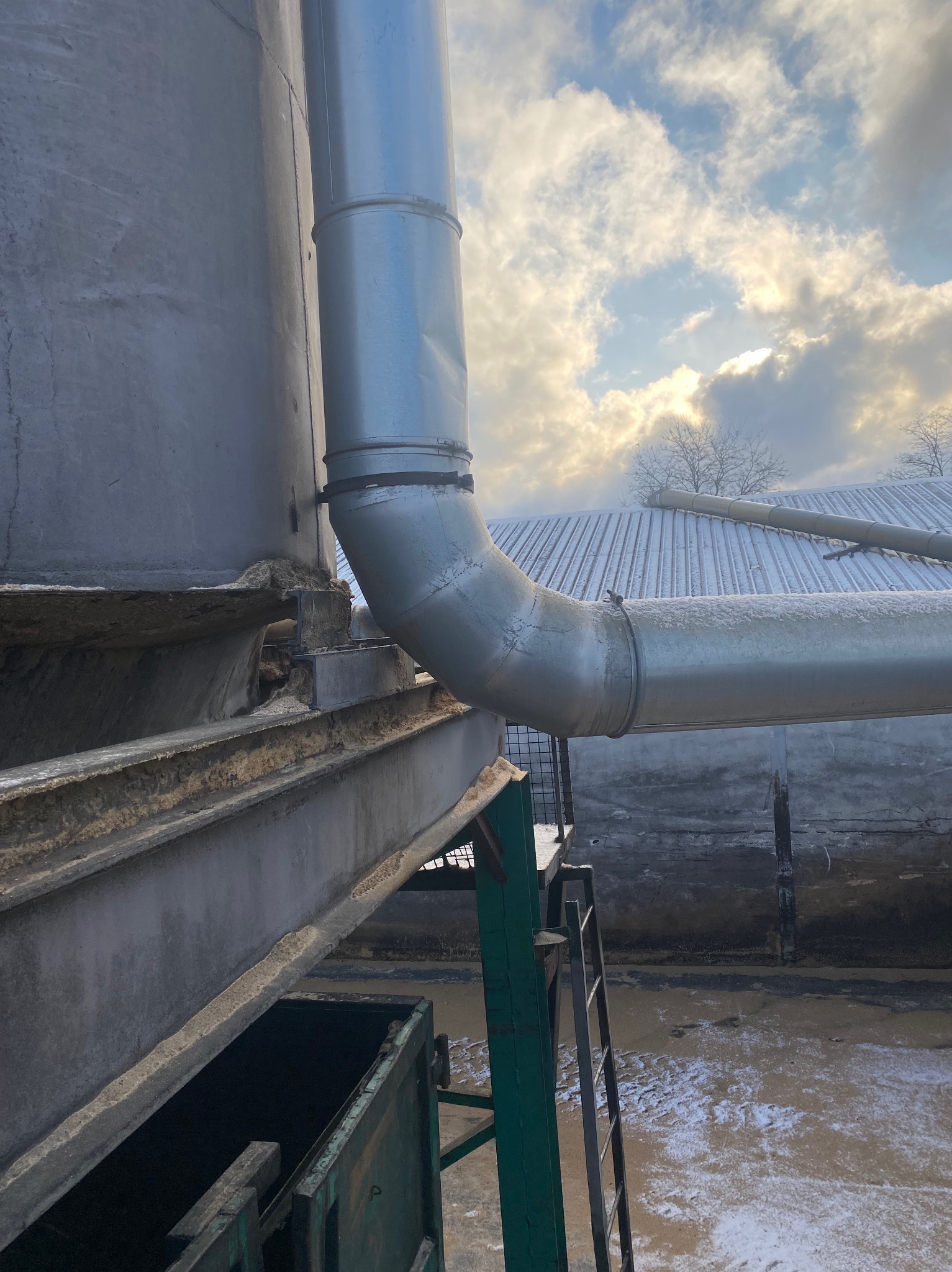 Metalowa rura przemysłowa z widocznymi spawami i elementami mocującymi, biegnąca wzdłuż zbiornika, na tle pochmurnego nieba i ośnieżonego dachu.