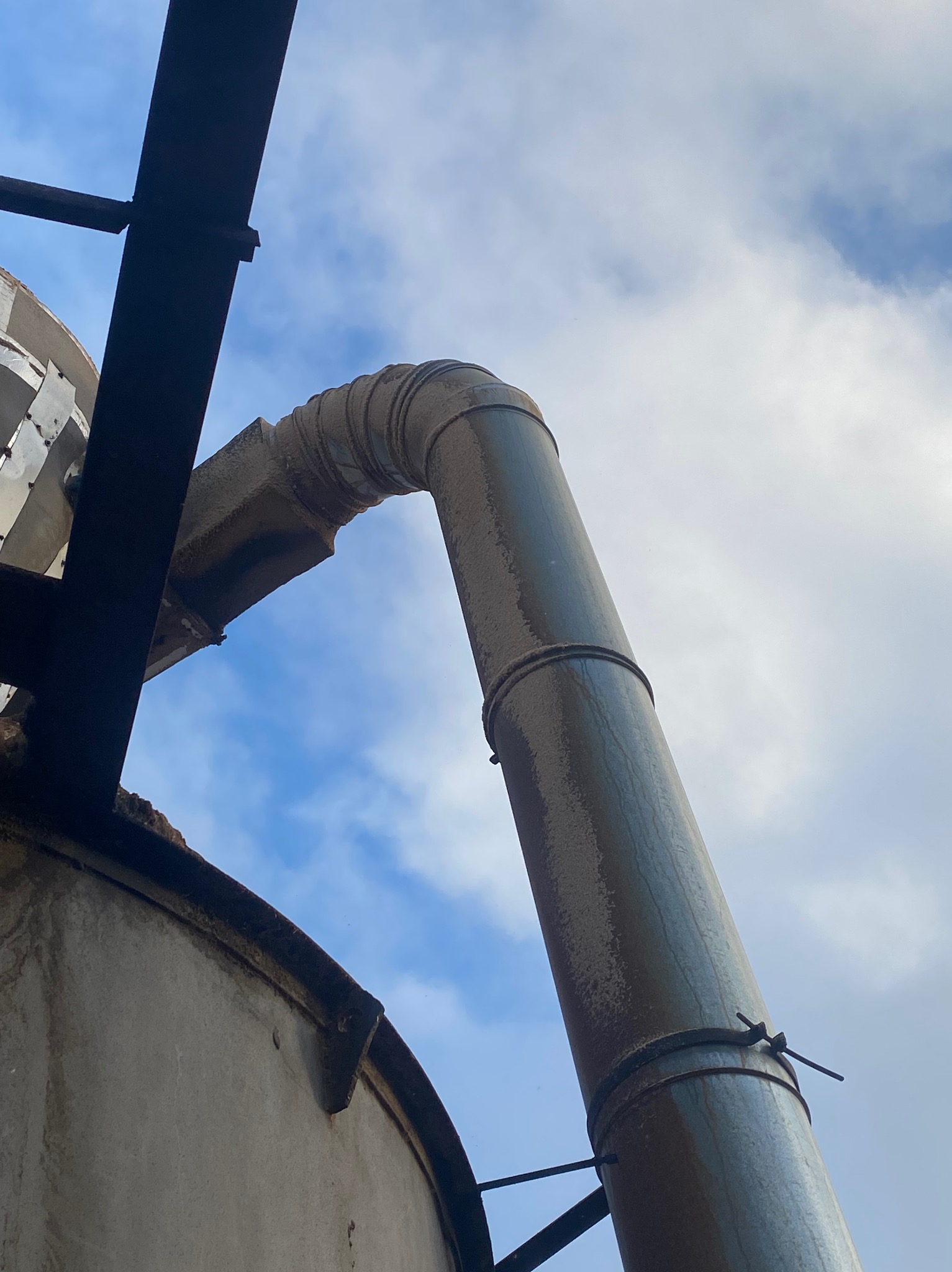 Metalowa konstrukcja z rurą w kształcie litery U, widok z dołu na tle nieba z chmurami. Widoczne elementy mocujące i spawane.