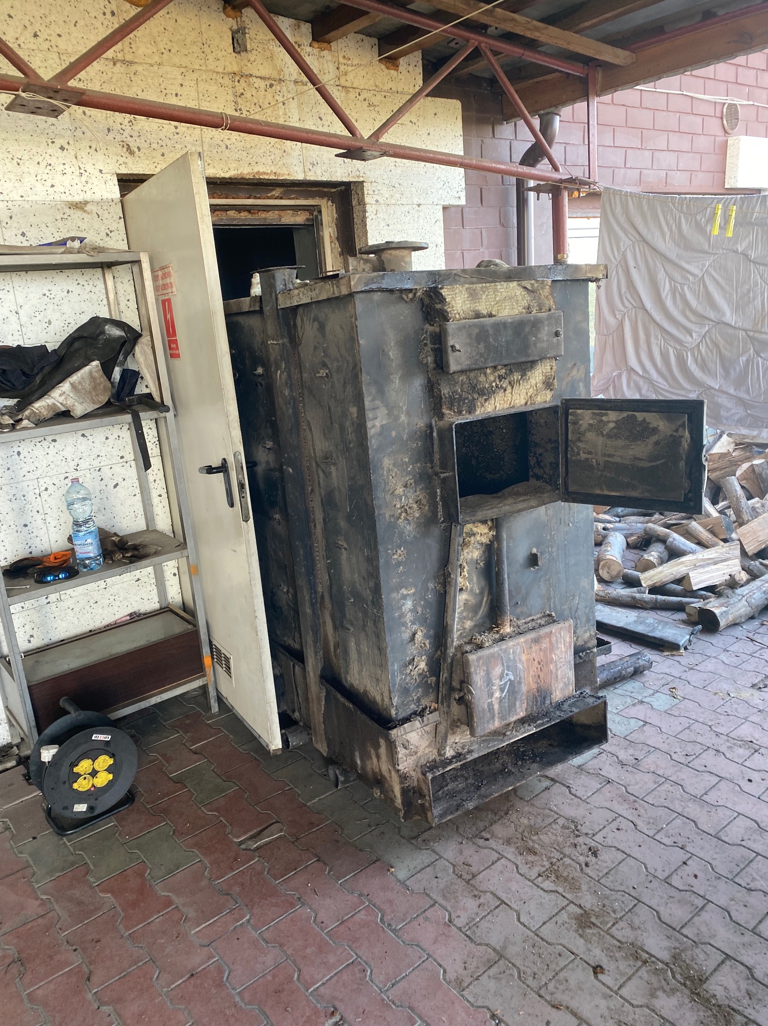 Stary, czarny, metalowy piec z otwartymi drzwiczkami, obok sterta drewna. W tle ściana z cegły i regał z narzędziami. Widoczne ślady użytkowania.
