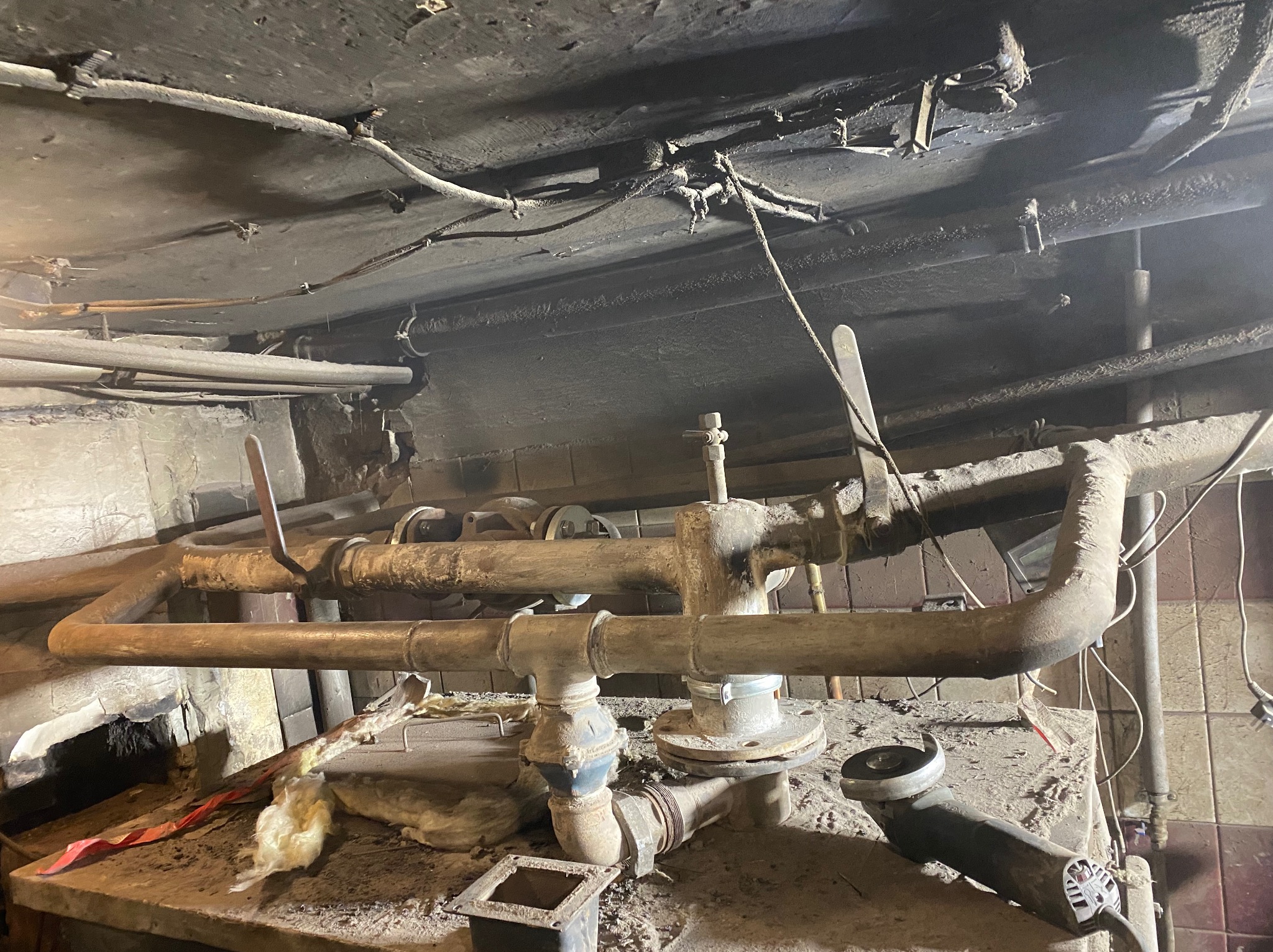 Stare, zardzewiałe rury w piwnicy, widoczne zawory i łączenia, zanieczyszczone otoczenie, w tle fragment ściany z płytkami i wiszące kable.