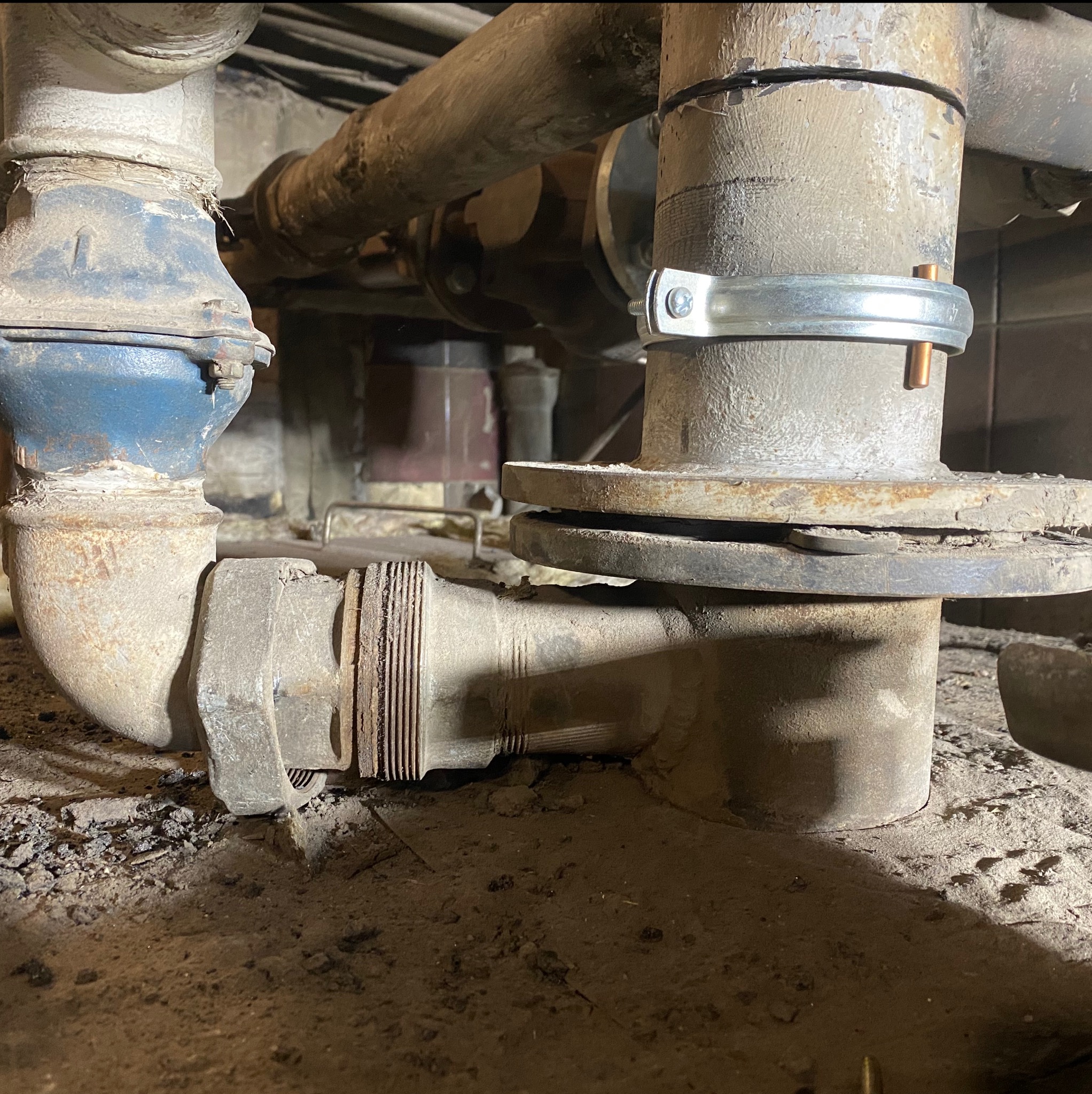 Szczegół złącza rur metalowych z widoczną korozją i śladami napraw, z bliska. Rury o różnych średnicach, połączone za pomocą złączek i obejm. Widoczny fragment podłoża.