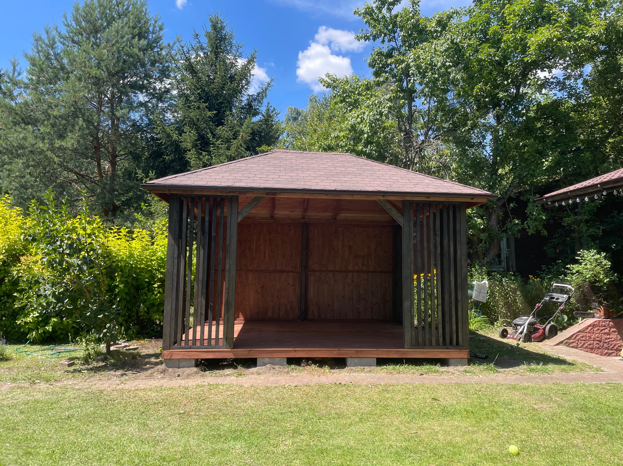 Drewniana altana z brązowym dachem na trawniku, otoczona zielenią. Widoczna kosiarka i piłka tenisowa. Słoneczny dzień.