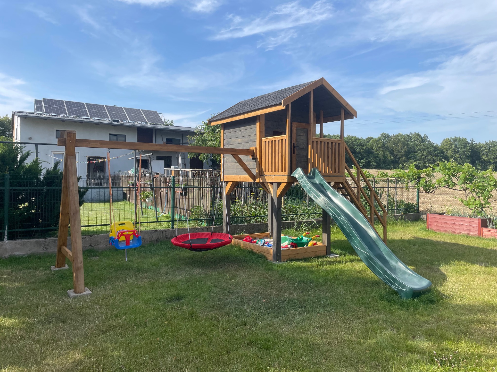 Drewniany plac zabaw z domkiem, zjeżdżalnią i huśtawką na trawniku. W tle dom z panelami słonecznymi i ogrodzenie. Całość w słoneczny dzień.