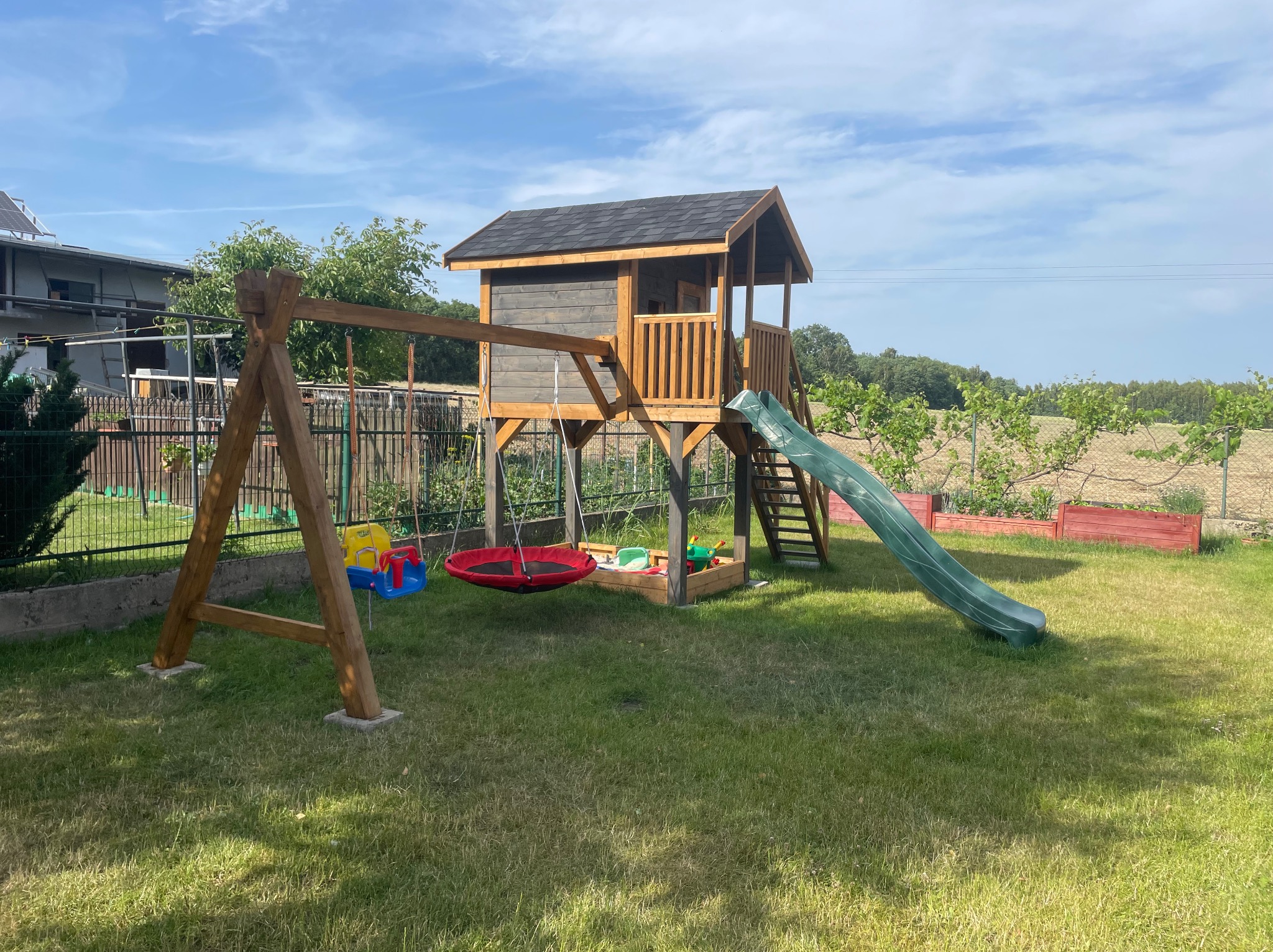 Drewniany plac zabaw z domkiem, zjeżdżalnią i huśtawkami na trawniku. Widok na ogród i budynek w tle, słoneczny dzień. Elementy placu pomalowane w kolorowe barwy.