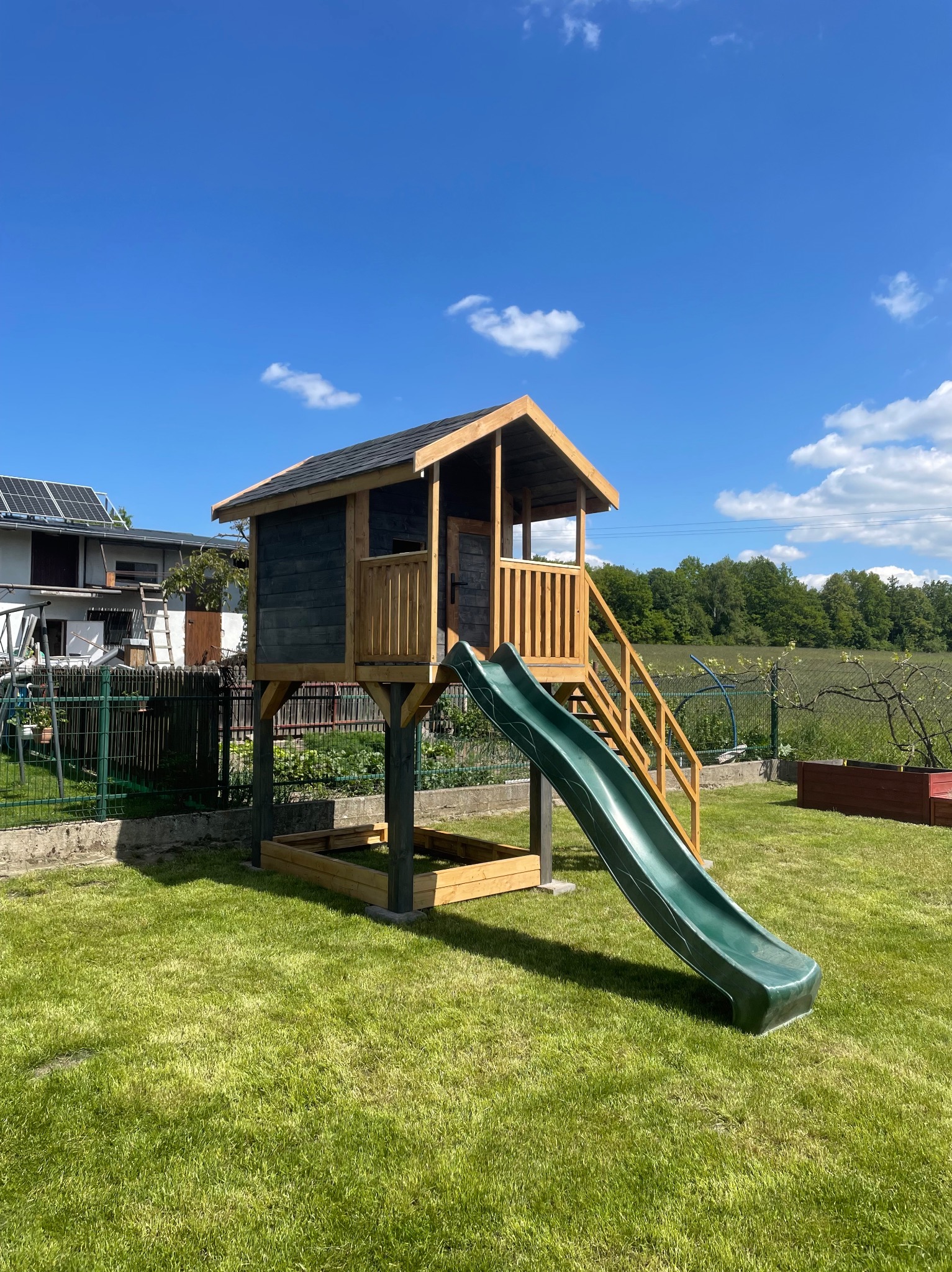 Drewniany domek dla dzieci z zieloną zjeżdżalnią, umieszczony na podwyższeniu, z piaskownicą w podstawie, na tle zadbanego trawnika i błękitnego nieba.