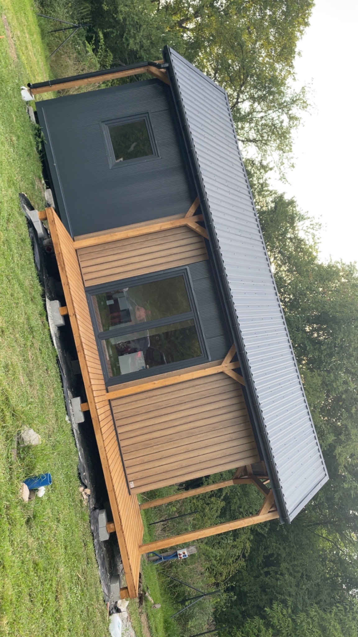Nowoczesna altana z drewnianą elewacją i metalowym dachem, widoczna z perspektywy trawnika. Uwagę przyciąga duże okno odbijające otoczenie.