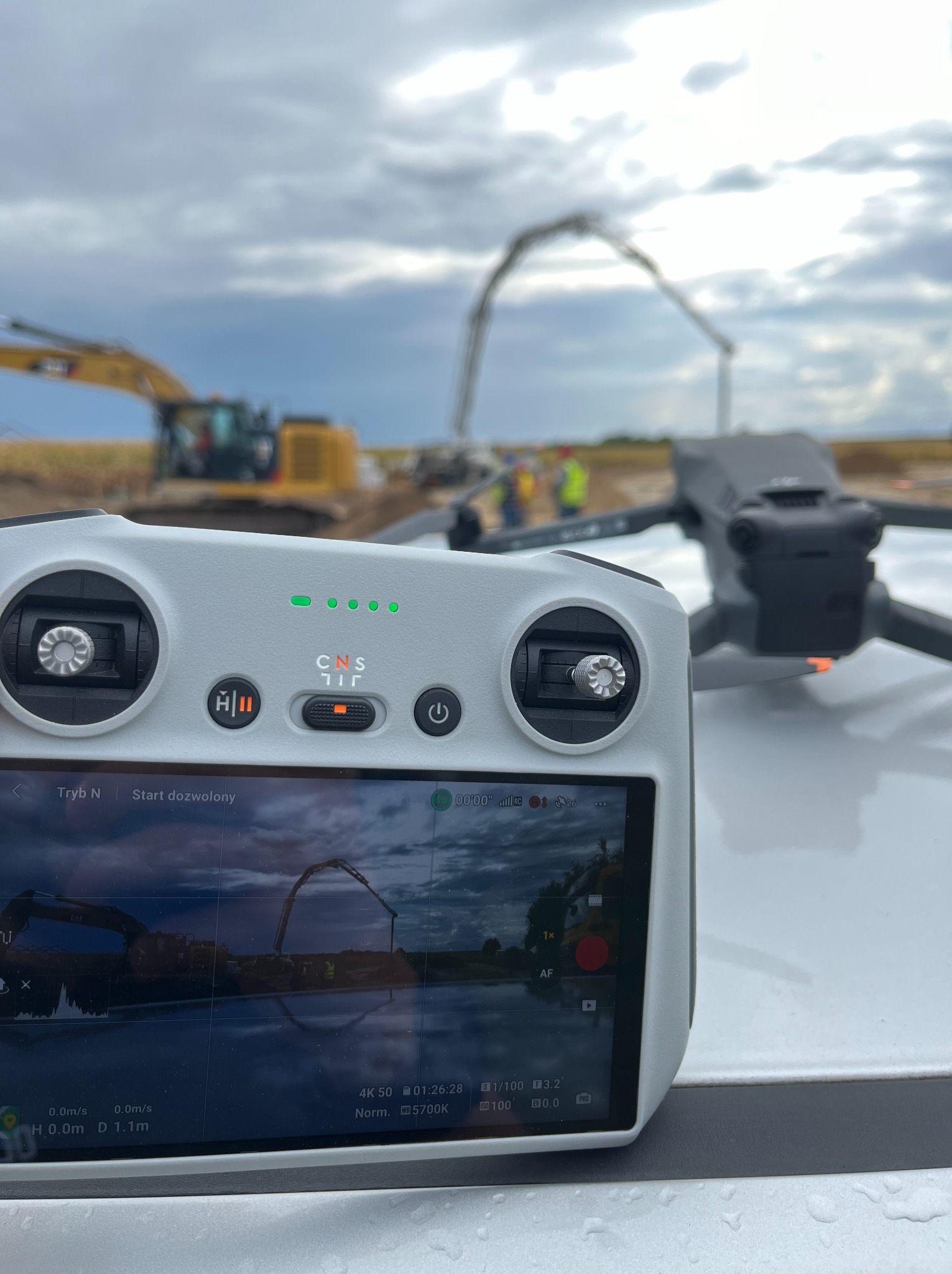 Dron DJI Mavic 3 z kontrolerem na dachu auta, w tle plac budowy z koparką i pompą do betonu. Ekran kontrolera pokazuje podgląd z drona. Pochmurne niebo.