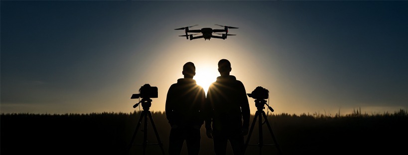 Sylwetki dwóch osób z kamerami na statywach i dronem na tle zachodzącego słońca. Kontrastowe ujęcie podkreśla sprzęt i osoby związane z filmowaniem.