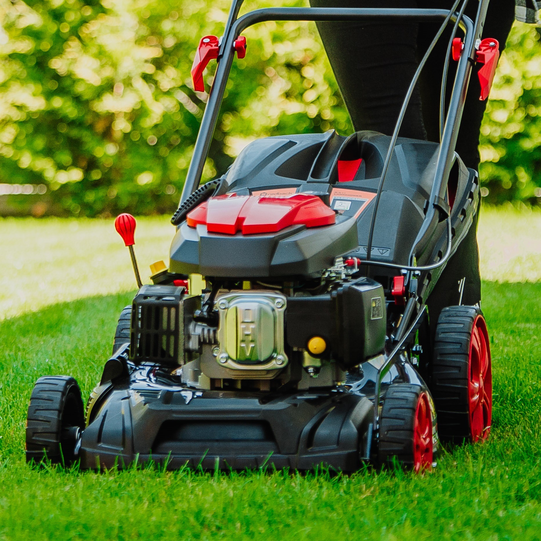 Czarno-czerwona kosiarka spalinowa na zielonej trawie, z widocznym silnikiem i elementami sterującymi. Lekko rozmyte tło z zielenią. Kosiarka gotowa do pracy.