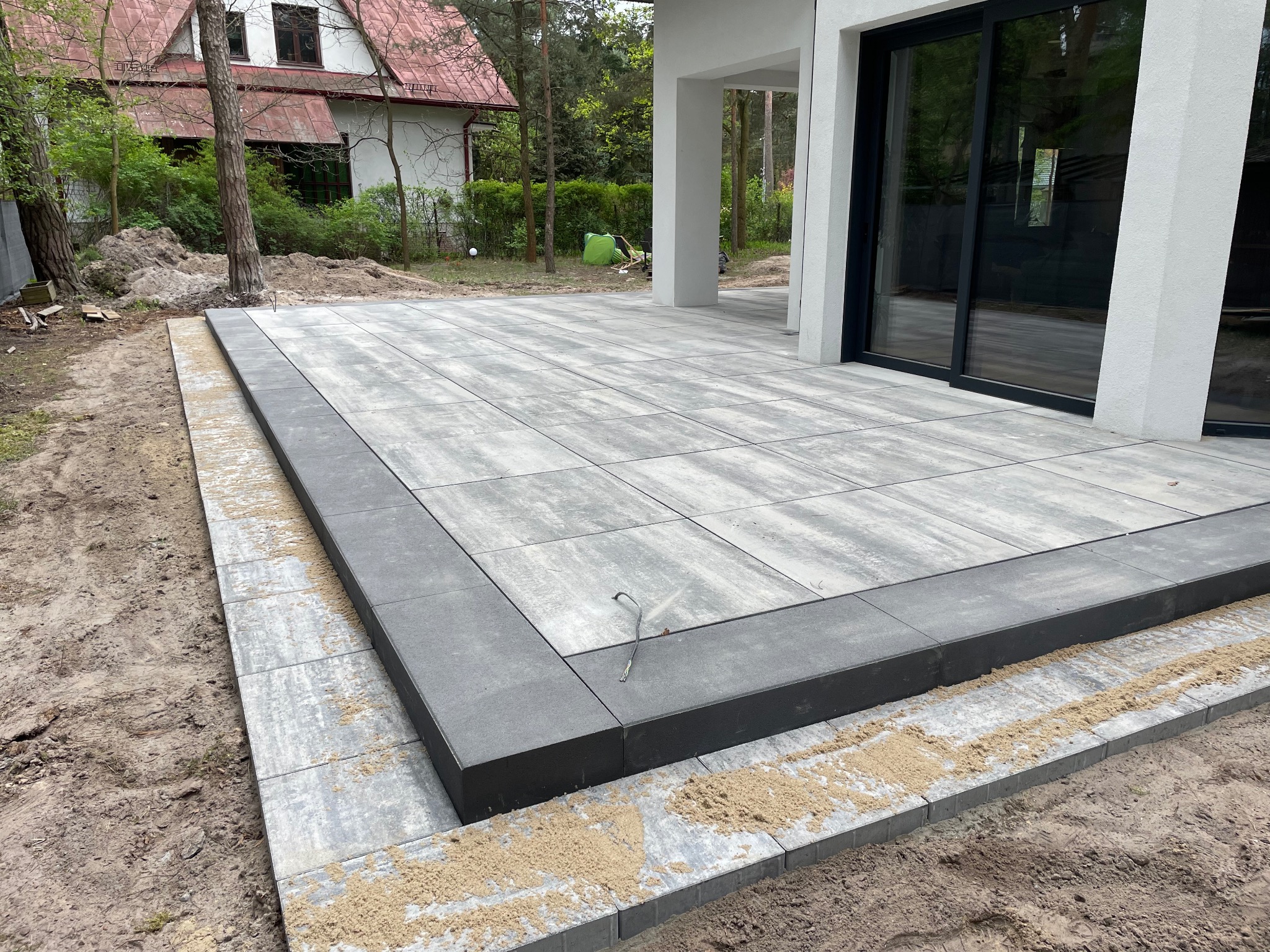 Nowoczesny taras z szarych płyt betonowych, obrzeża z ciemnego betonu, widok z boku, Warszawa. Świeżo ułożona nawierzchnia przy domu z dużymi oknami.