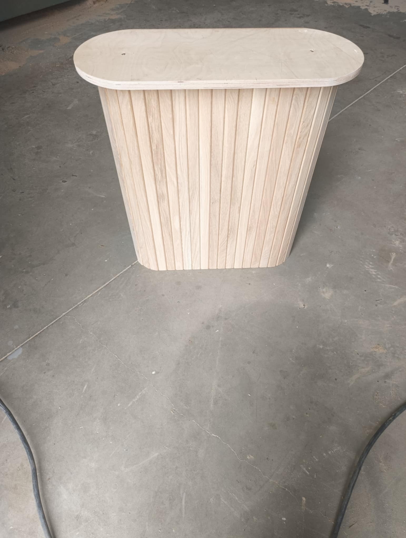 Drewniana szafka nocna z pionowymi listwami i owalnym blatem z jasnego drewna na szarym betonowym podłożu. Minimalistyczny design, stolarka na wymiar.