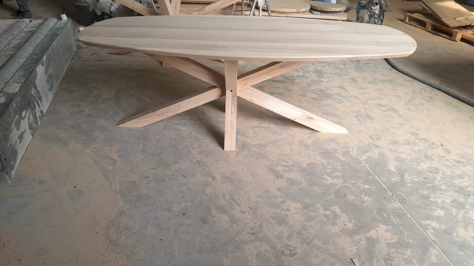 Owalny, surowy stół z jasnego drewna na oryginalnej, krzyżowej podstawie. Widoczne ślady obróbki, warsztatowy klimat. Blat gładki, nielakierowany.