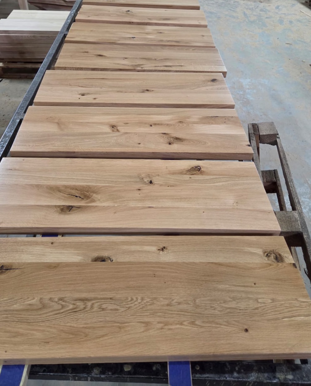 Jasne, dębowe blaty drewniane ułożone równolegle na metalowym regale, ukazujące naturalne słoje i unikalne detale drewna. Widoczne palety w tle.