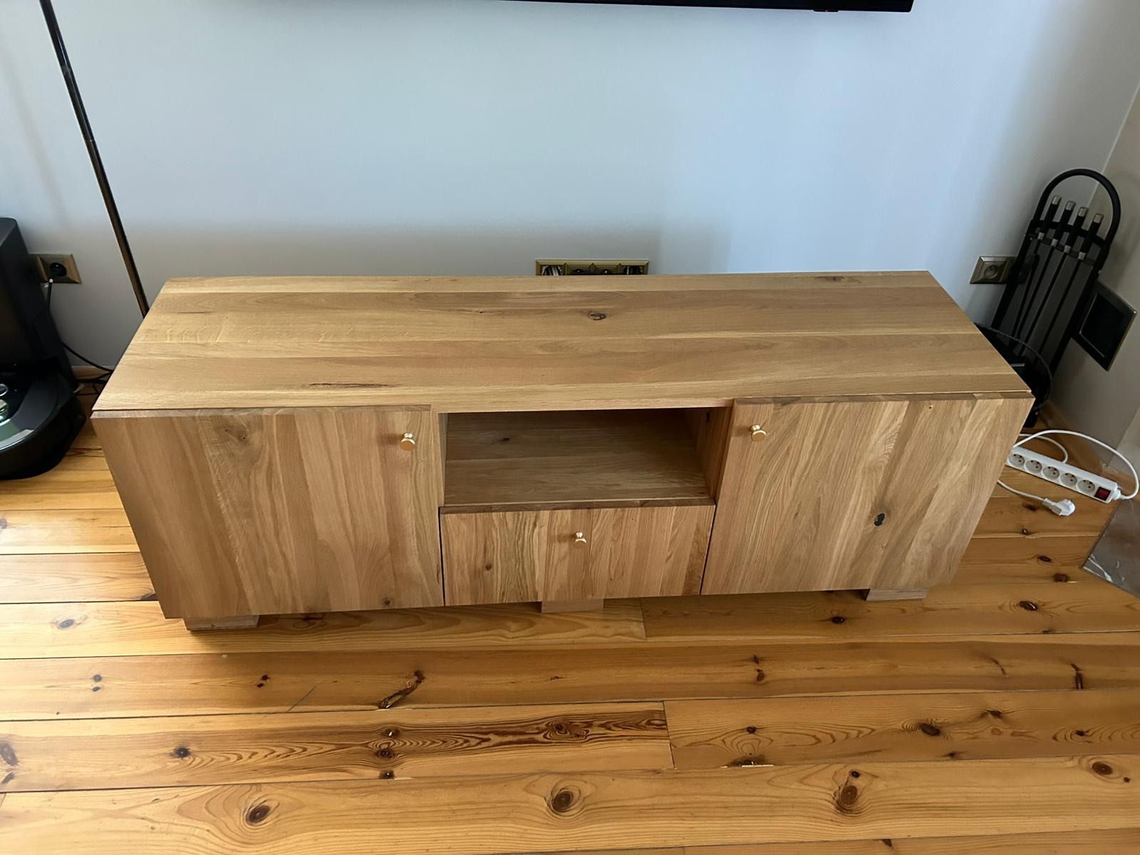 Drewniana szafka RTV z szufladą i szafkami po bokach, wykonana na wymiar, stoi na parkiecie. Widoczne złote gałki i naturalne usłojenie drewna.