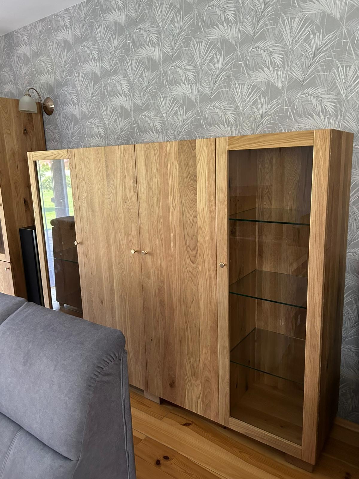 Drewniana szafa z przeszklonymi drzwiami i półkami, widoczna na tle ściany w liściasty wzór. Jasne drewno, minimalistyczne uchwyty, podłoga z desek.