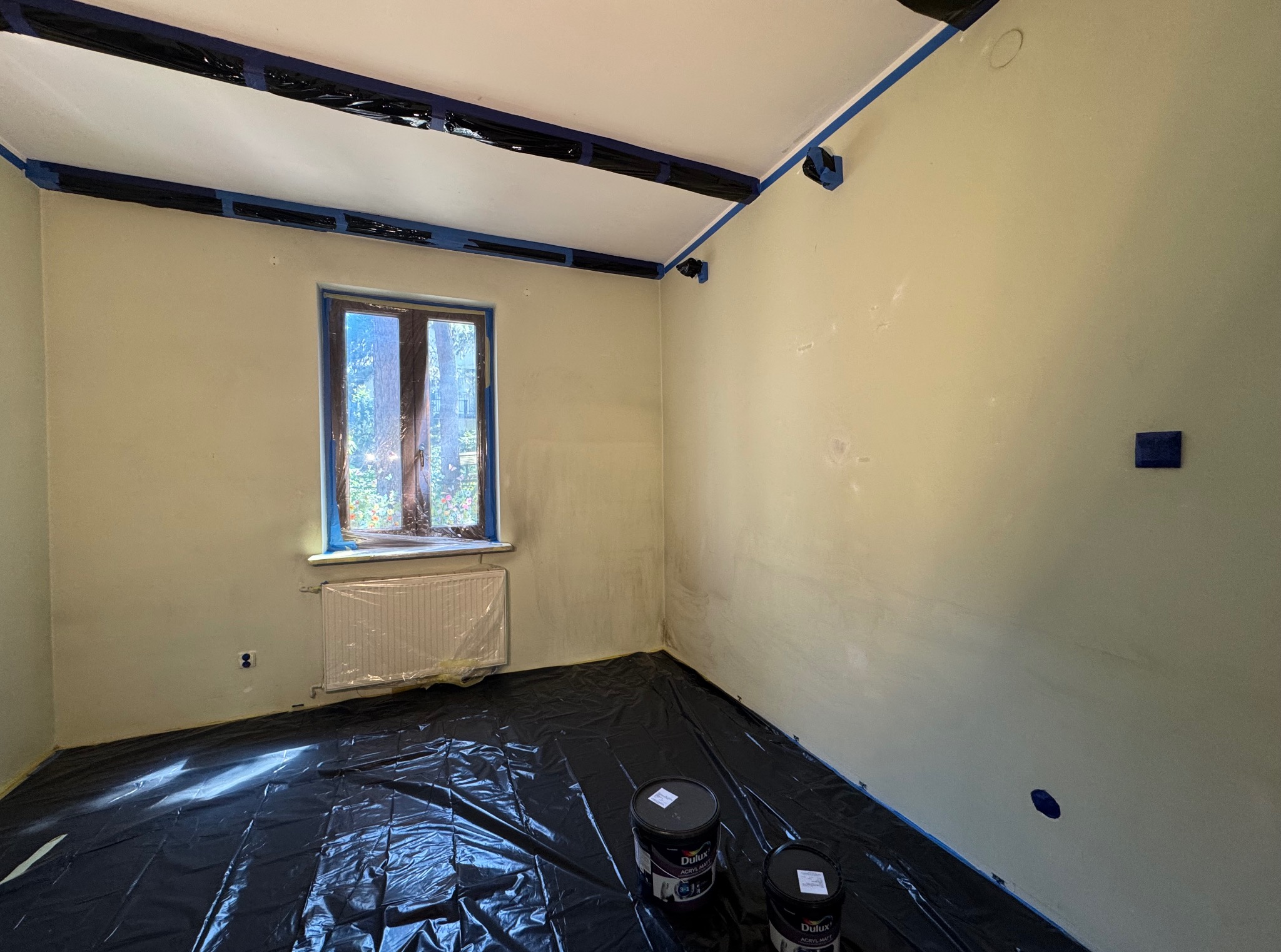 Pokój przygotowany do malowania: folia ochronna na podłodze, taśma malarska na listwach i oknach, widoczne dwa wiadra z farbą Dulux.