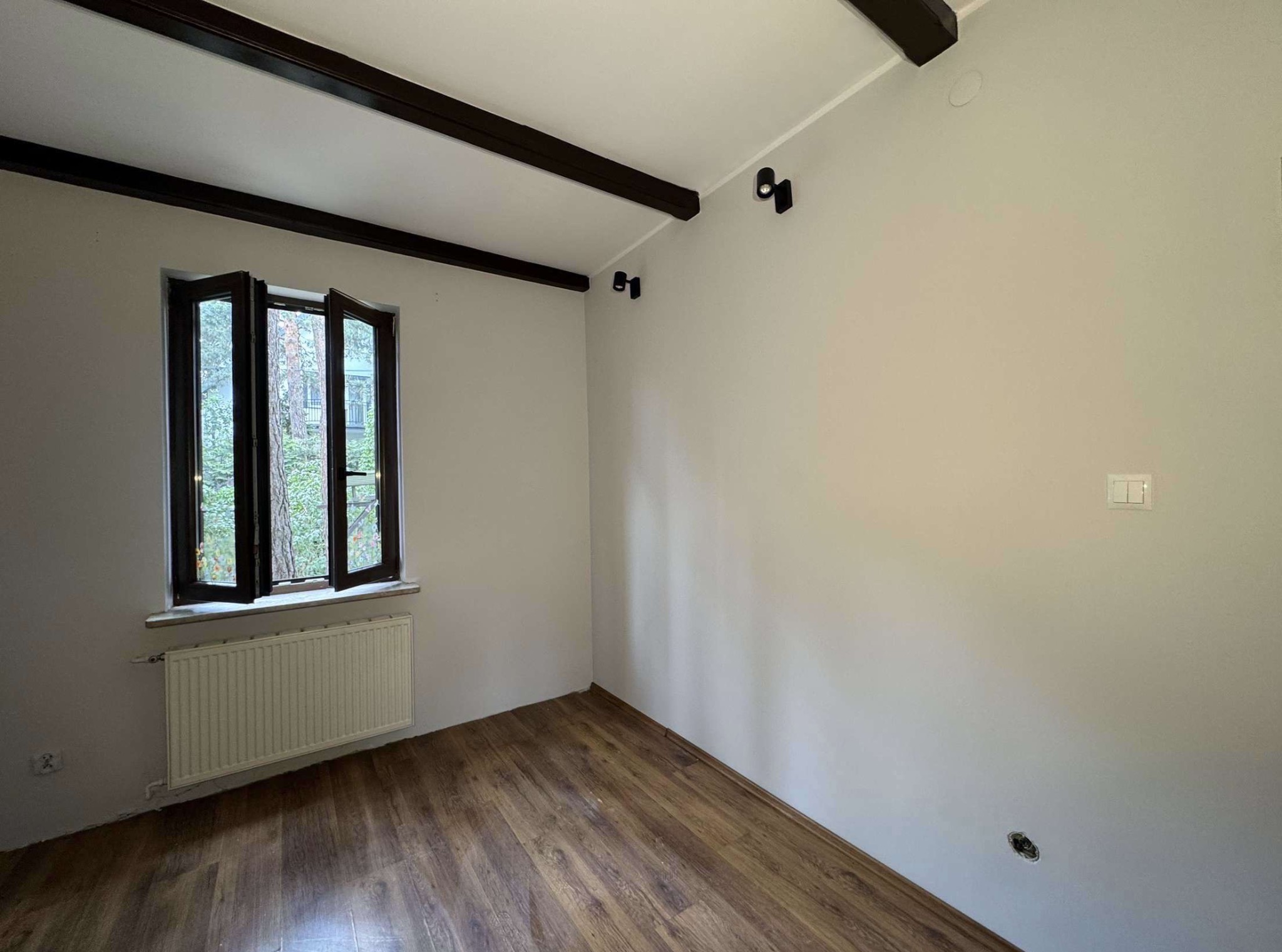 Jasne wnętrze po remoncie: pomalowane ściany, drewniana podłoga, okno z widokiem na zieleń, ciemne belki sufitowe i czarne lampy. Gładkie ściany gotowe do aranżacji.