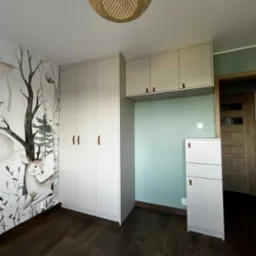 Pok&oacute;j dziecięcy z szafą na wymiar w jasnej kolorystyce, z drewnianymi uchwytami i ozdobną tapetą z motywem zwierząt. Całość dopełnia drewniana podłoga i lampa.