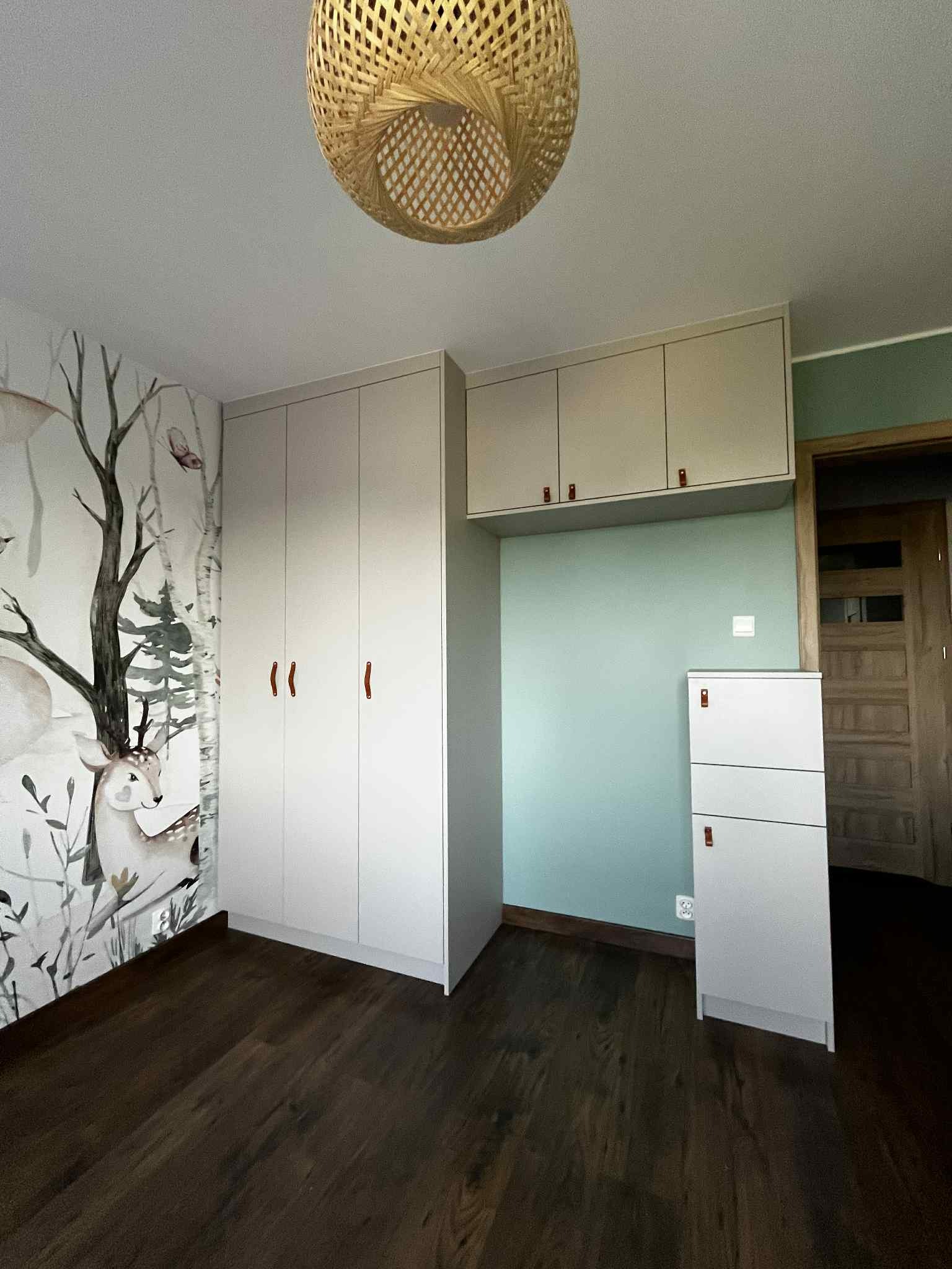 Pokój dziecięcy z szafą na wymiar w jasnej kolorystyce, z drewnianymi uchwytami i ozdobną tapetą z motywem zwierząt. Całość dopełnia drewniana podłoga i lampa.