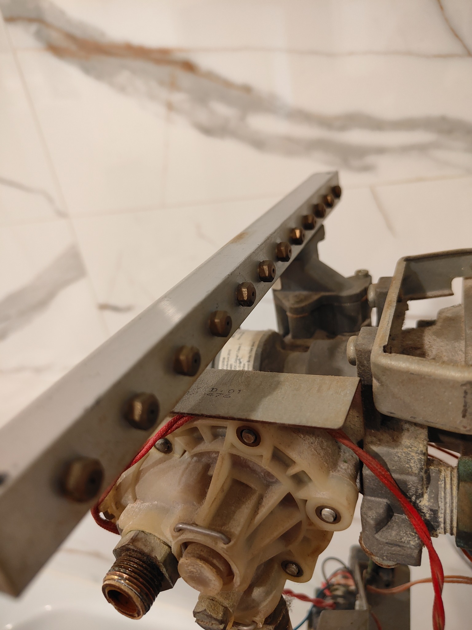 Demontaż elementu pieca gazowego z widocznymi śrubami, przewodami i zabrudzeniami, prawdopodobnie w trakcie serwisu na tle marmurowej ściany.