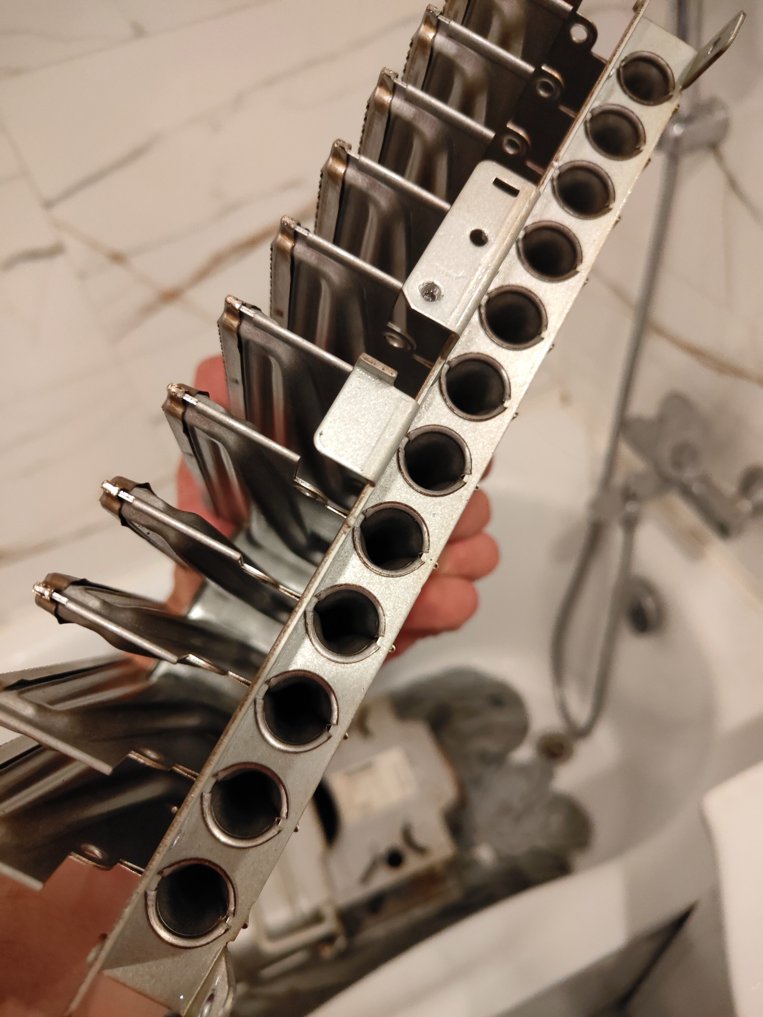 Wyjęty z pieca gazowego, metalowy palnik z rzędami otworów, trzymany w dłoni na tle łazienki. Widoczne ślady użytkowania, wnętrze palnika z osadem.