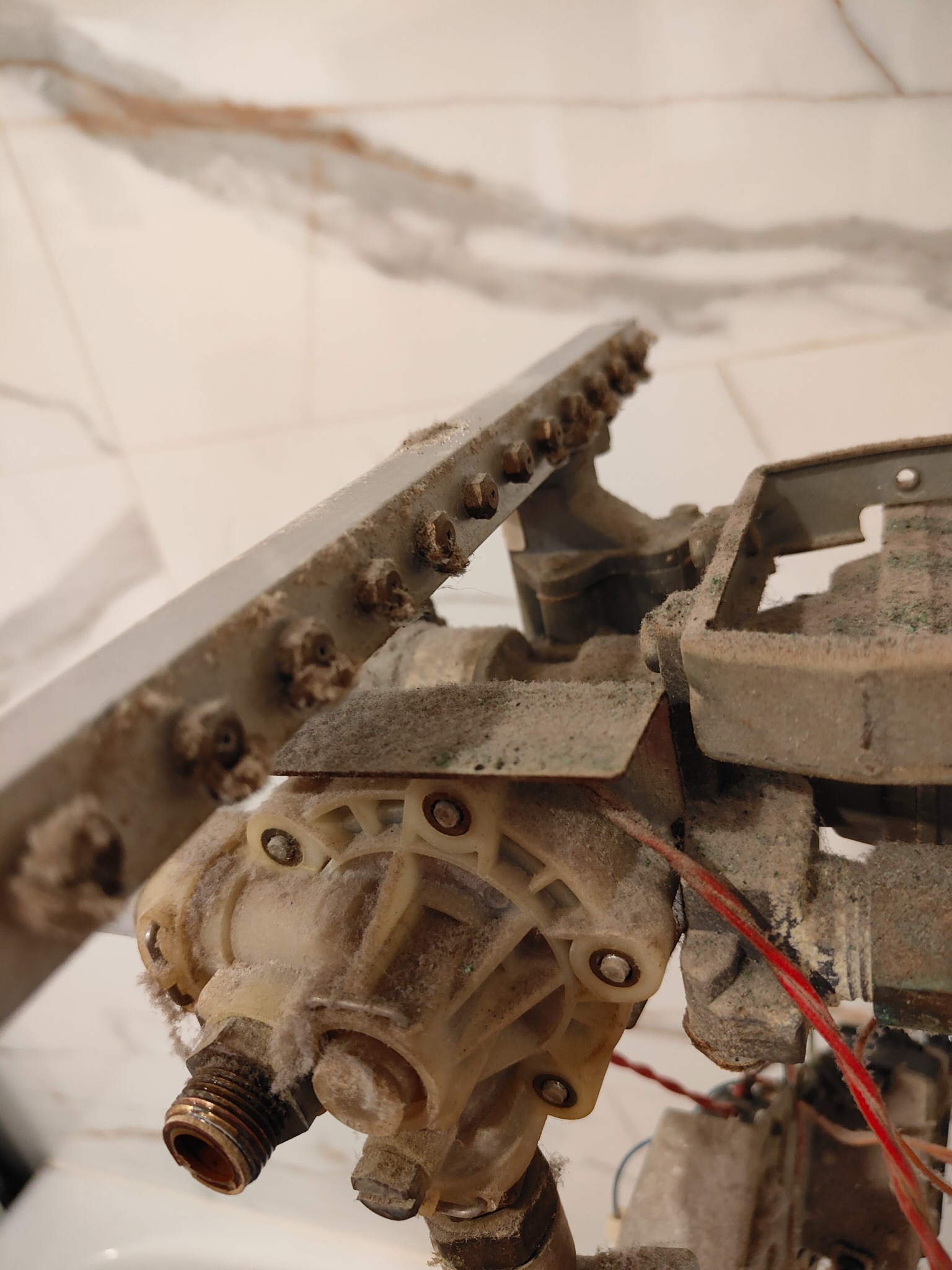 Zdemontowany, bardzo zakurzony element pieca gazowego z widocznymi przewodami, złączkami i elementami konstrukcyjnymi na tle białej ściany z marmurowym wzorem.