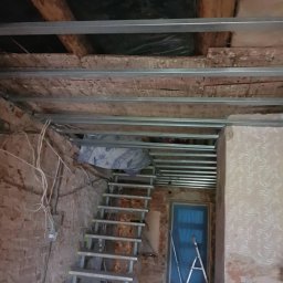 Cezary Grzegorz - Montaż metalowej konstrukcji sufitu podwieszanego, widoczne profile i stare drewniane elementy. W tle schody i drabina oparta o niebieskie drzwi.