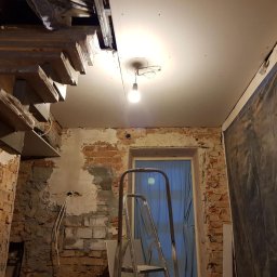 Cezary Grzegorz - Wnętrze w trakcie remontu: surowa cegła, częściowo otynkowana ściana, zamontowana płyta karton-gips na suficie, schody z drewnianymi stopniami w folii ochronnej i drabina.
