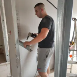 Pracownik w trakcie montażu profili do płyt karton-gips za pomocą specjalistycznych nożyc, widoczne narzędzia i fragment konstrukcji ściany działowej.