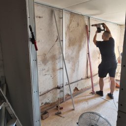 Cezary Grzegorz - Prace przygotowawcze do montażu płyty karton-gips: pracownik wierci w metalowym profilu na ścianie. Widoczne narzędzia i materiały budowlane.