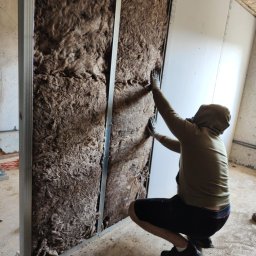 Cezary Grzegorz - Pracownik w trakcie montażu izolacji z wełny mineralnej w metalowym stelażu pod płytę gipsowo-kartonową na poddaszu. Widoczne elementy konstrukcyjne dachu.