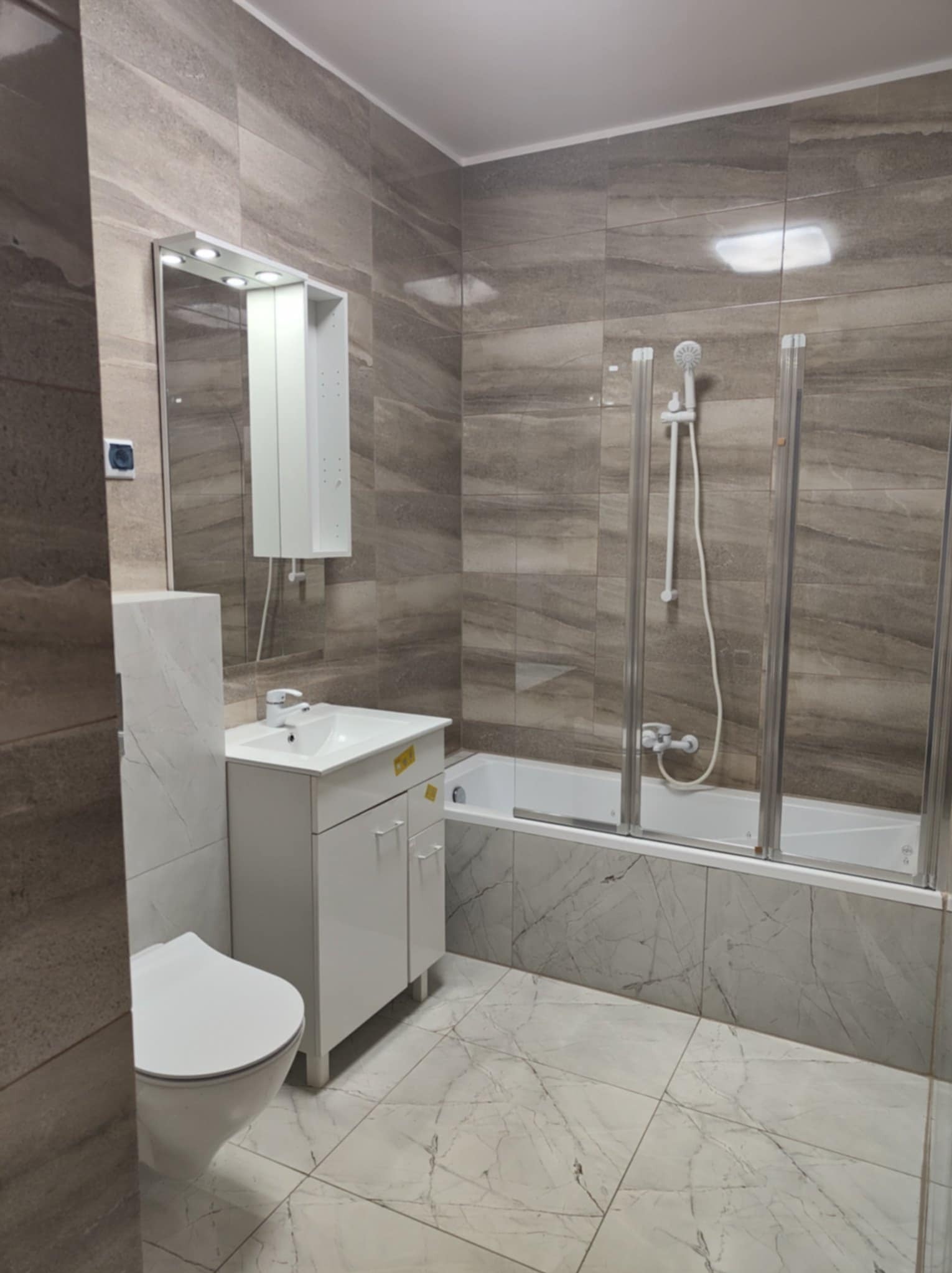 Wykończona łazienka z wanną z parawanem, umywalką z szafką i toaletą. Płytki imitujące marmur na ścianach i podłodze. Nowoczesne oświetlenie lustra.