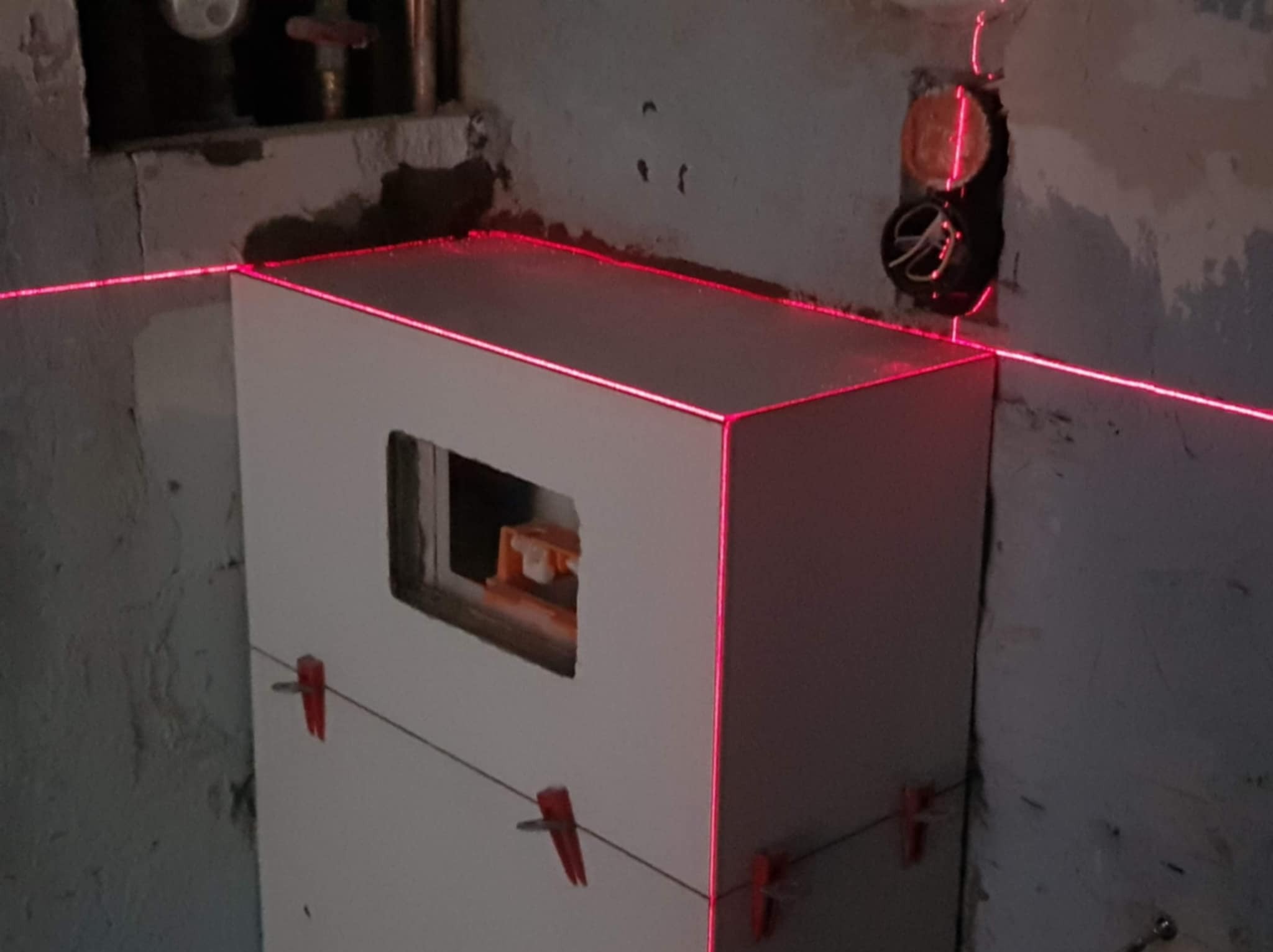 Biała obudowa stelaża WC w trakcie montażu, poziomowana laserowo. Widoczne klipsy glazurnicze i surowa ściana łazienki. Prace wykończeniowe w toku.