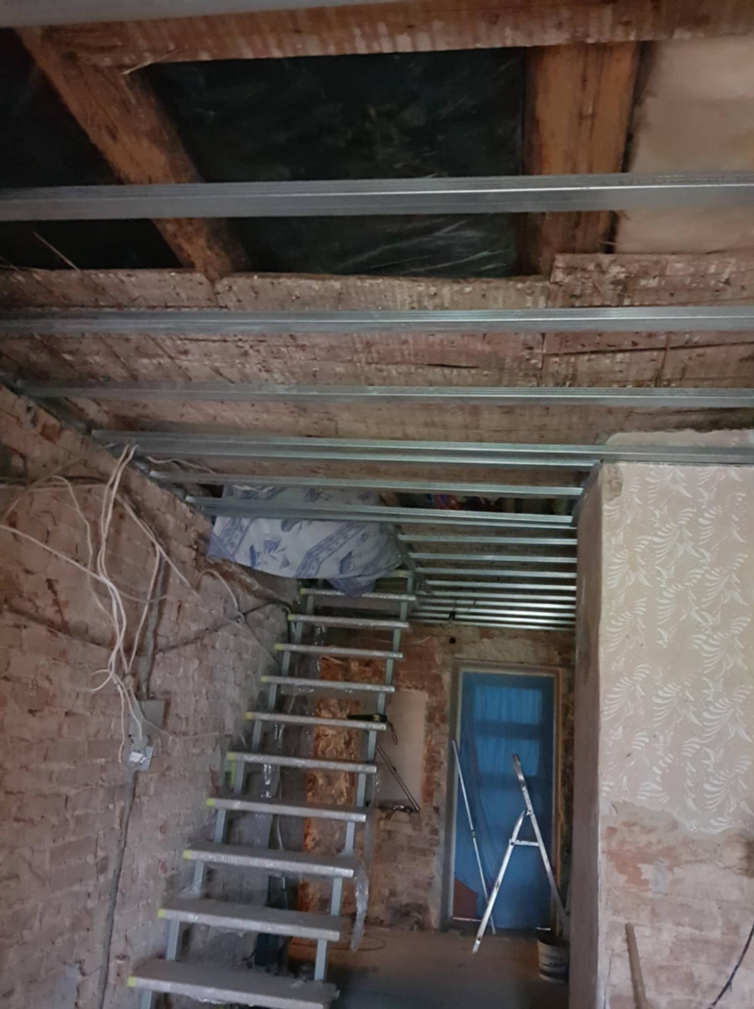 Montaż metalowej konstrukcji sufitu podwieszanego, widoczne profile i stare drewniane elementy. W tle schody i drabina oparta o niebieskie drzwi.