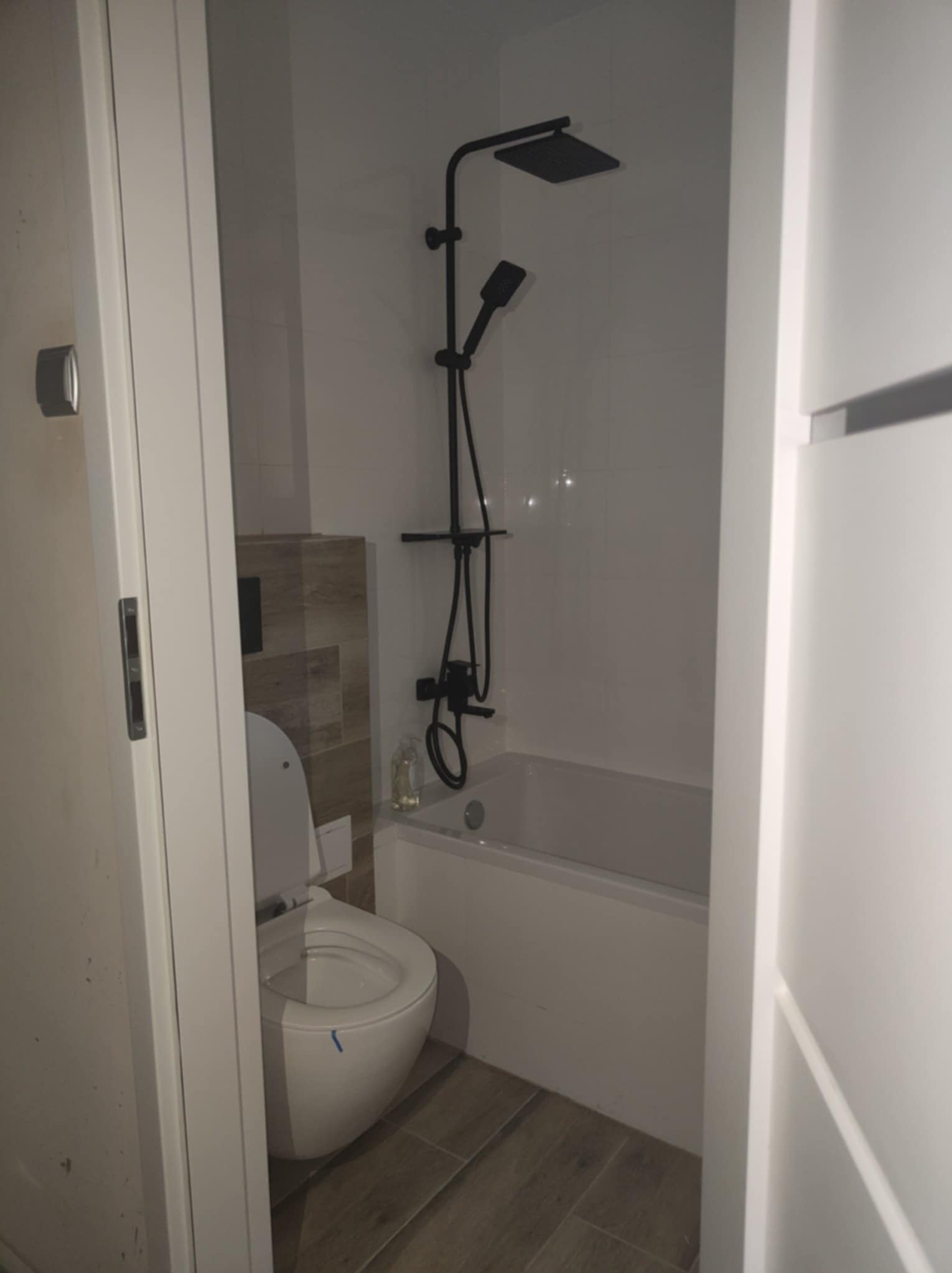 Nowoczesna łazienka w bieli i drewnie: WC, wanna z czarną armaturą, płytki imitujące drewno. Minimalistyczny design, widok zza uchylonych drzwi.