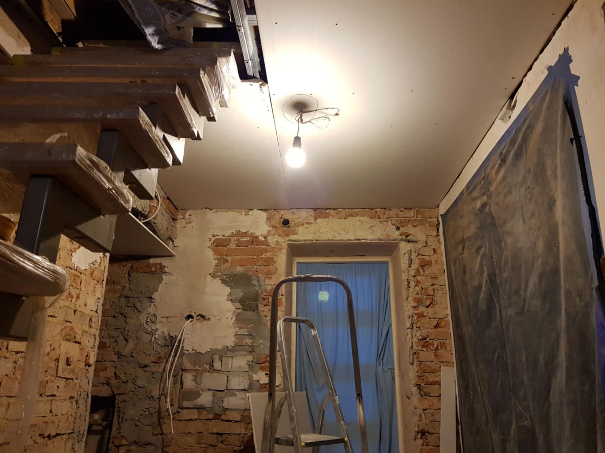Wnętrze w trakcie remontu: surowa cegła, częściowo otynkowana ściana, zamontowana płyta karton-gips na suficie, schody z drewnianymi stopniami w folii ochronnej i drabina.
