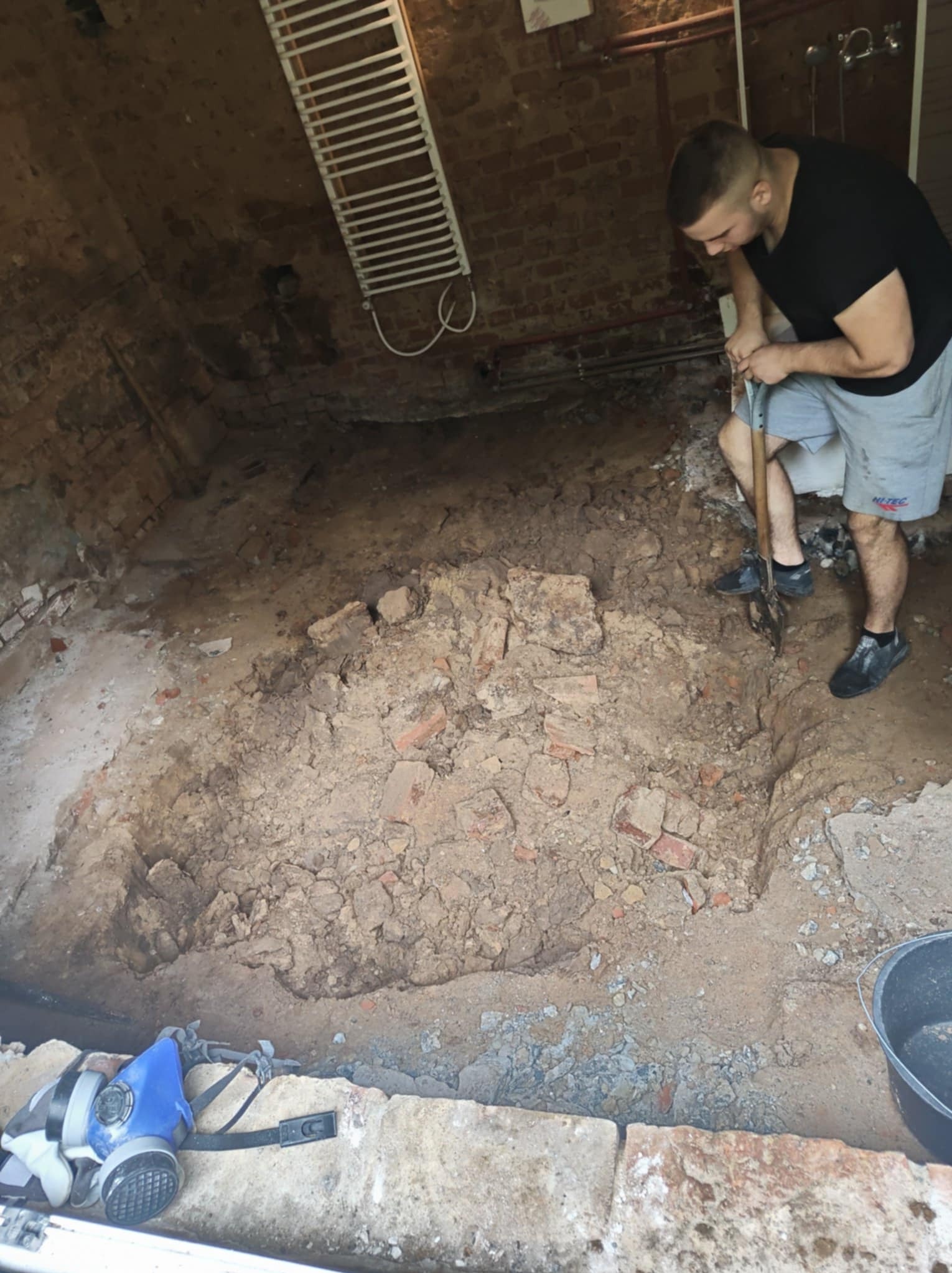 Demontaż podłogi w starej łazience z ceglanymi ścianami. Mężczyzna w szortach kopie łopatą. Widoczny grzejnik drabinkowy i maska ochronna.