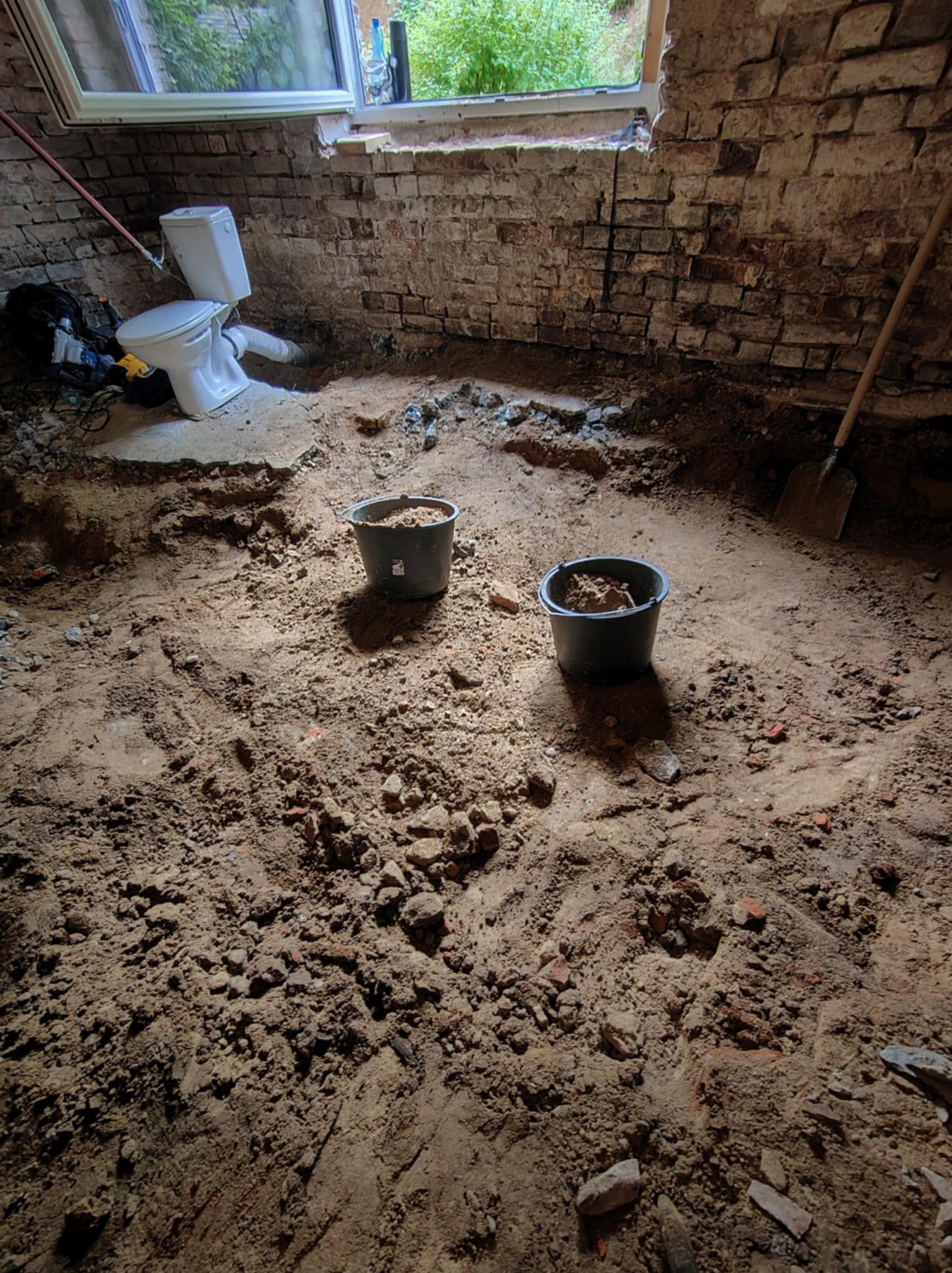Wnętrze w trakcie remontu łazienki: widoczne ceglane ściany, wykopana podłoga z dwoma wiadrami ziemi, odłączona toaleta i okno z widokiem na zieleń.