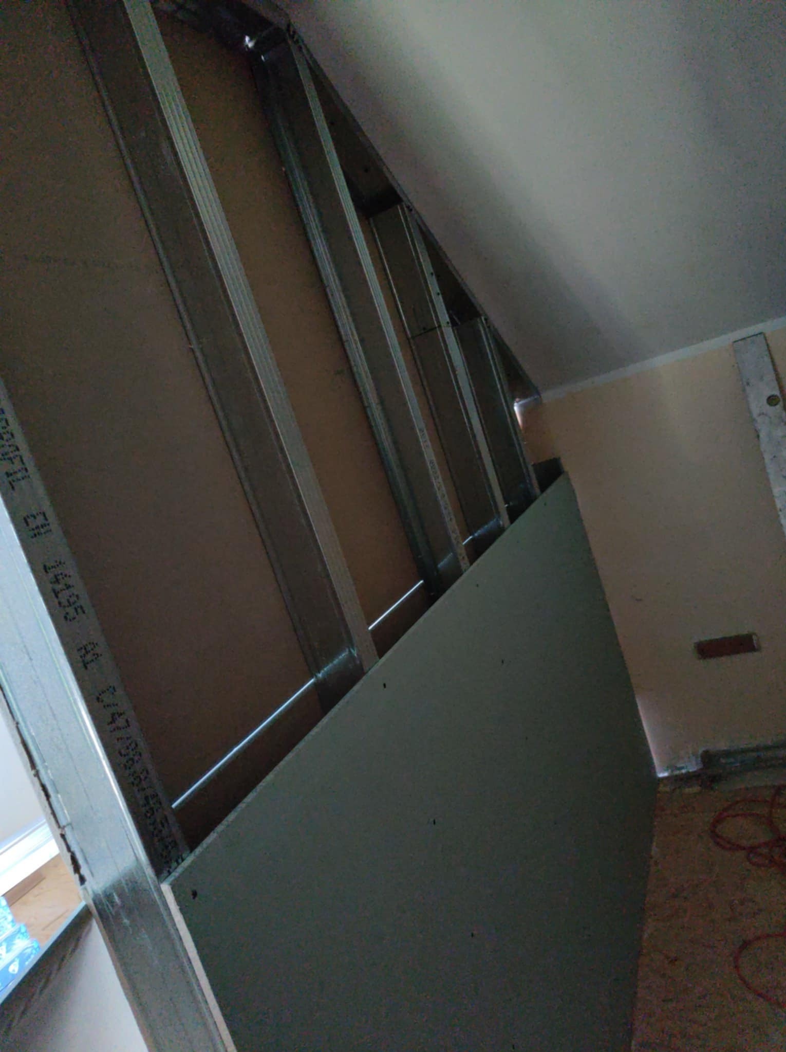 Skośna ściana z płyt karton-gips, widoczne metalowe profile konstrukcyjne. Częściowo zamontowana płyta. Ujęcie z dołu, blisko ściany, z widocznym fragmentem podłogi.
