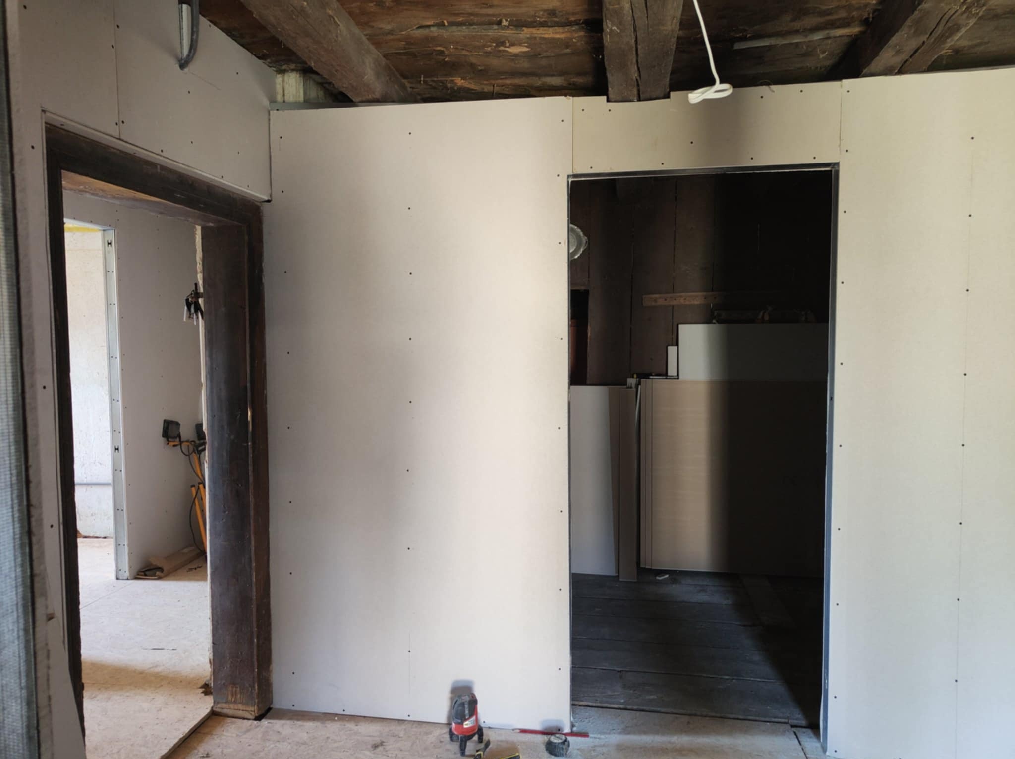 Montaż płyt kartonowo-gipsowych w trakcie remontu. Widoczne drewniane belki stropowe i surowe, drewniane ościeżnice drzwiowe.