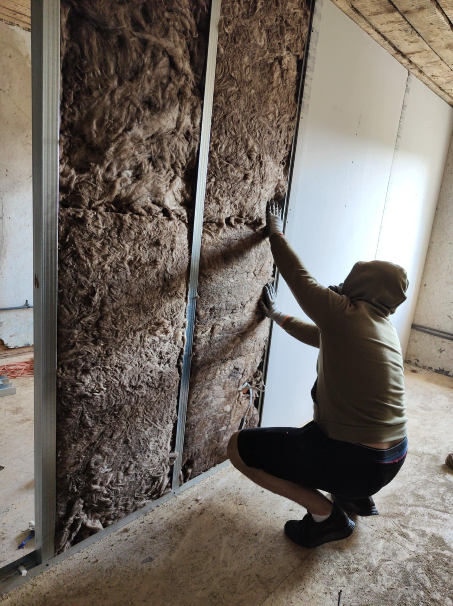 Pracownik w trakcie montażu izolacji z wełny mineralnej w metalowym stelażu pod płytę gipsowo-kartonową na poddaszu. Widoczne elementy konstrukcyjne dachu.