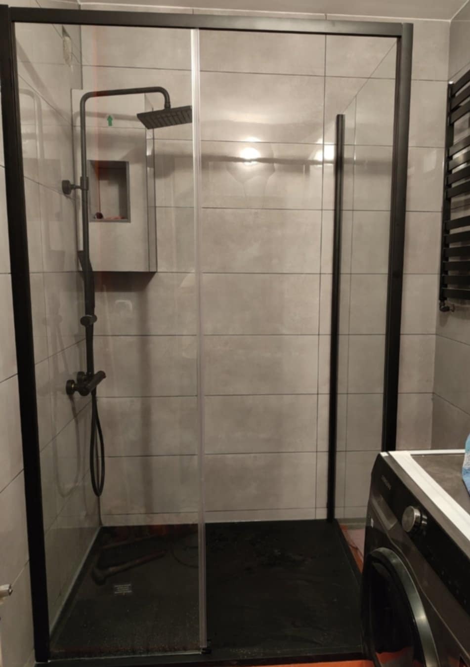 Nowoczesna kabina prysznicowa z czarnymi profilami, szklane drzwi przesuwne, szare płytki na ścianach, czarny brodzik. Minimalistyczny design łazienki.