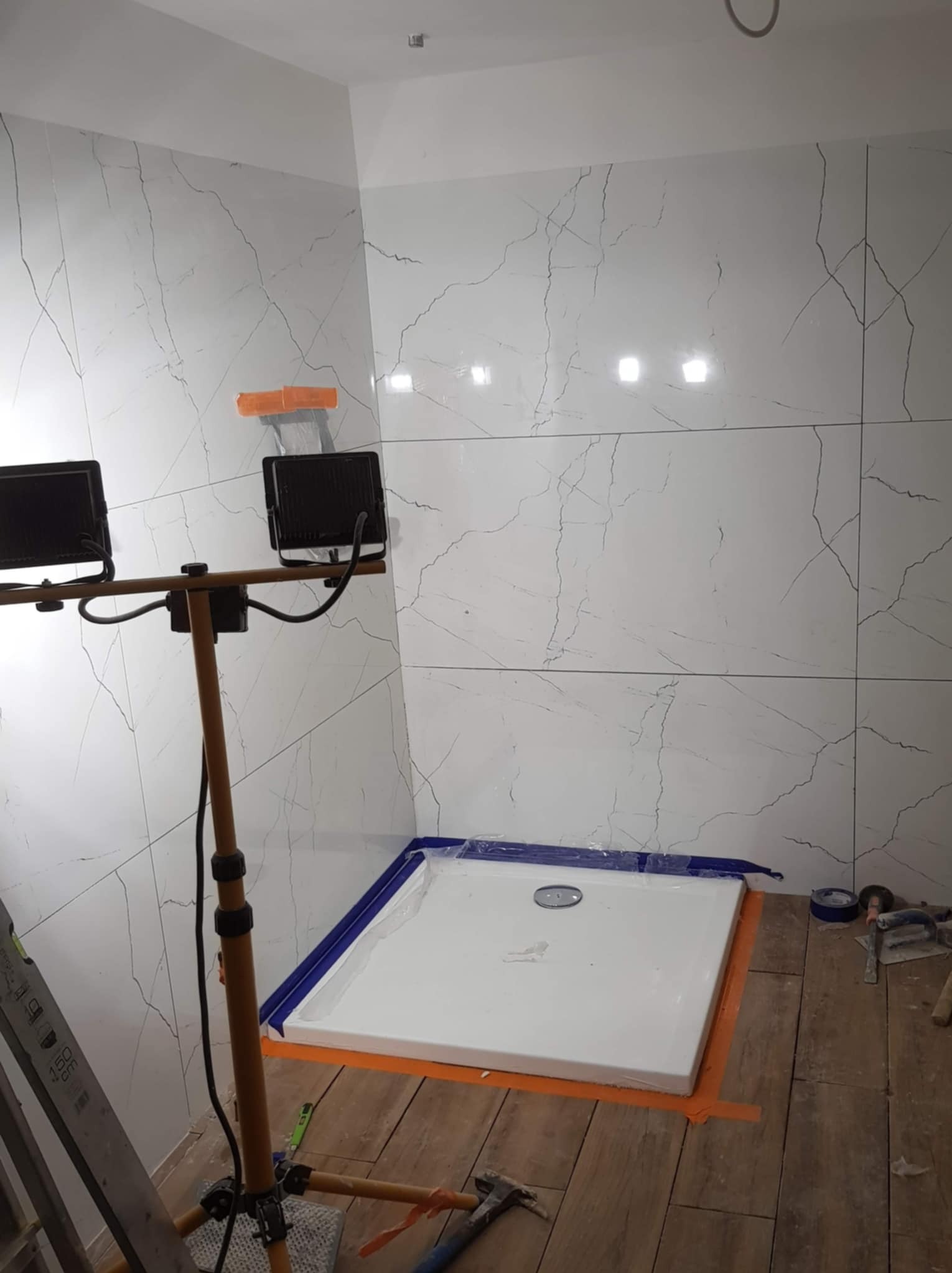 Białe, marmurowe płytki na ścianie łazienki, zamontowany brodzik zabezpieczony folią, oświetlenie budowlane. Widoczne narzędzia glazurnicze.