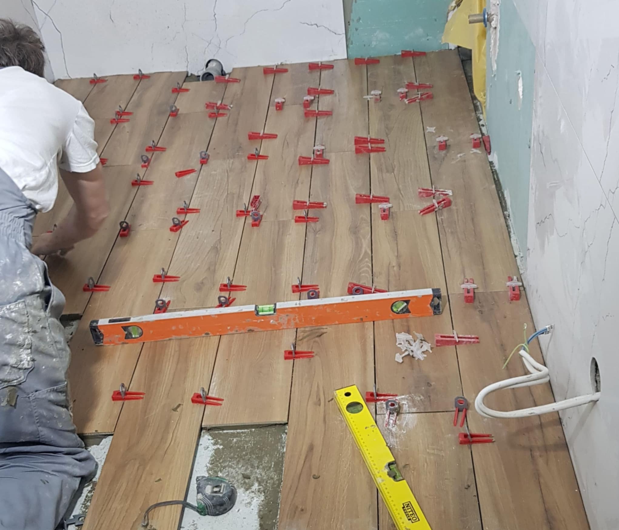 Układanie drewnopodobnych płytek na podłodze z użyciem poziomicy i czerwonych klipsów poziomujących. Widoczny fragment ściany z instalacją elektryczną.