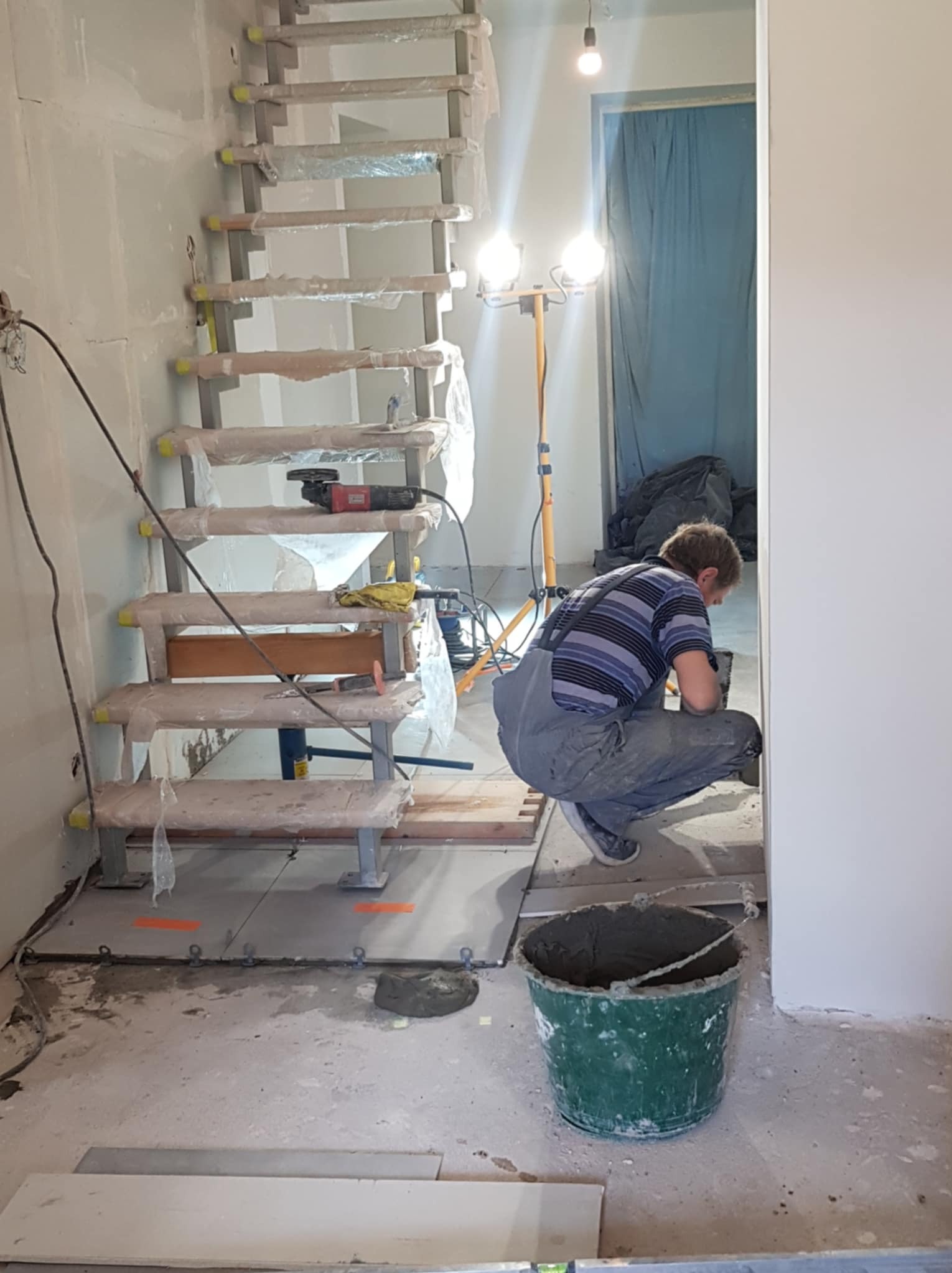 Prace wykończeniowe: glazurnik w trakcie układania płytek pod schodami, widoczne narzędzia, wiadro z zaprawą i zabezpieczone schody. Hala w stanie surowym.