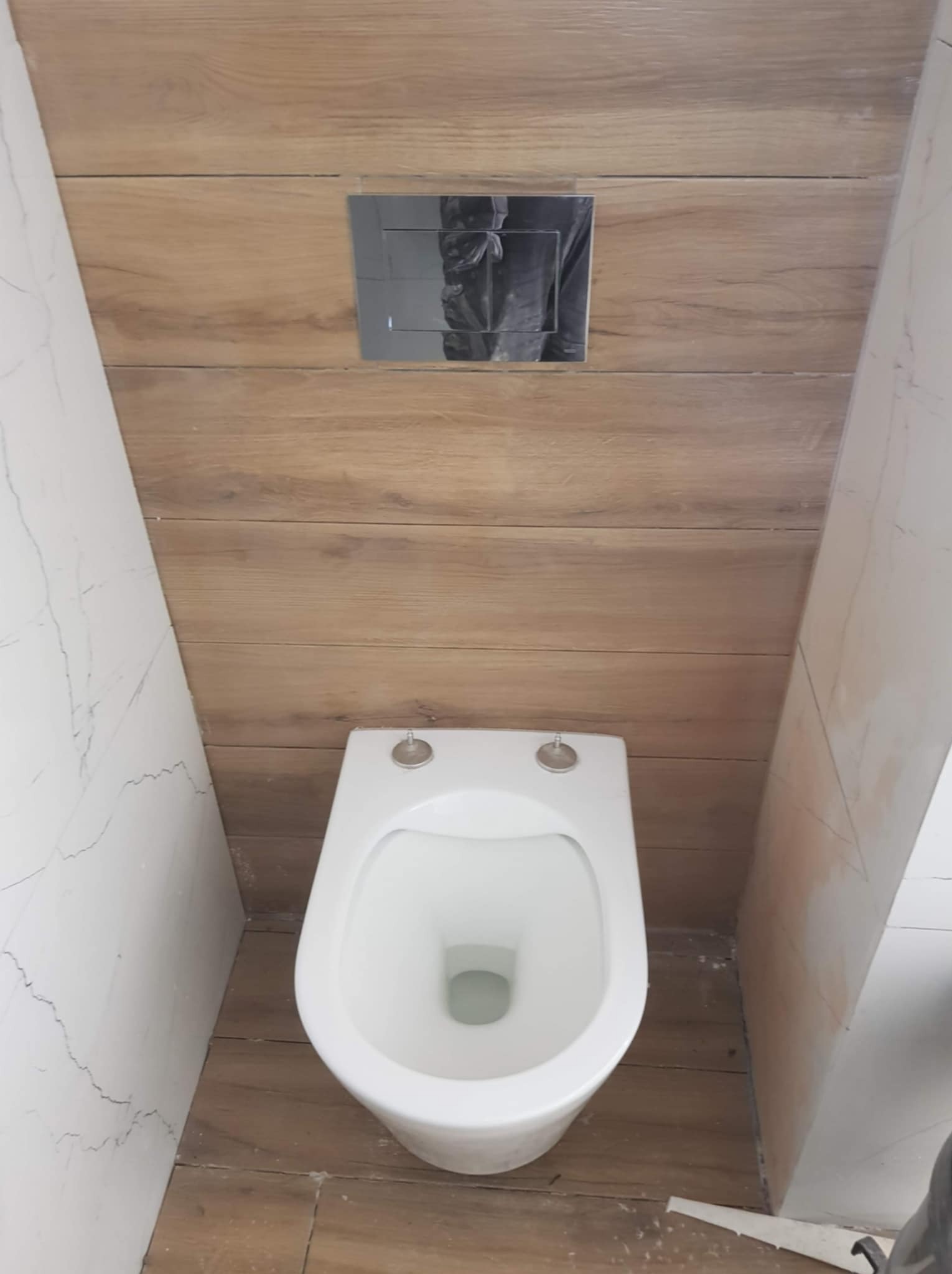 Zamontowana toaleta wisząca z chromowanym przyciskiem spłukującym na ścianie wyłożonej płytkami imitującymi drewno, w łazience z marmurowymi płytkami.