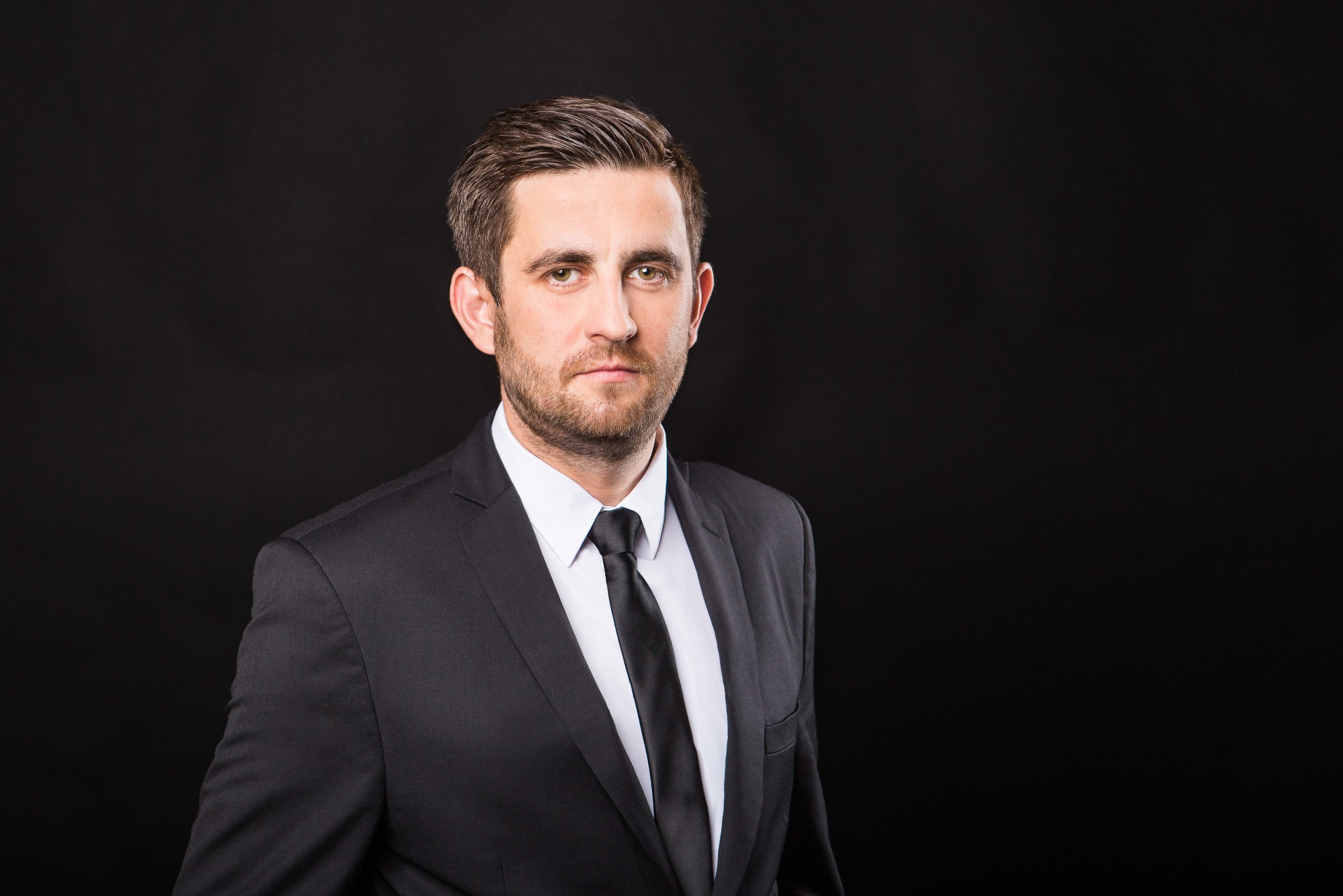 Portret biznesowy mężczyzny w czarnym garniturze i białej koszuli na ciemnym tle. Powaga i profesjonalizm w spojrzeniu.