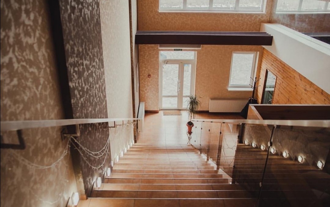 Jasne wnętrze: widok ze schodów na hol z rośliną i oknami. Ściany wykończone w beżowych odcieniach, z elementami dekoracyjnymi. Widoczny fragment drewnianej boazerii.