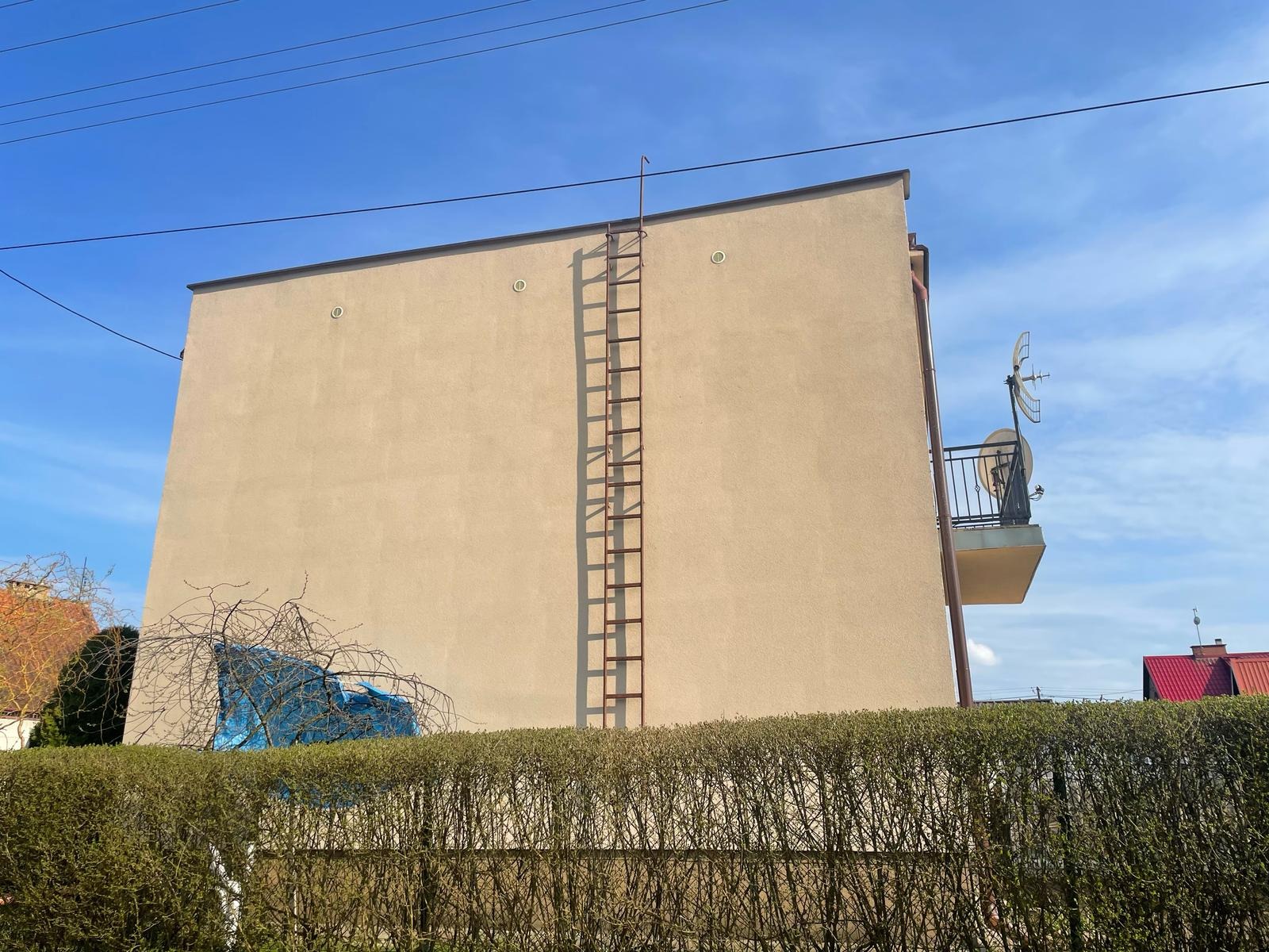 Elewacja budynku z beżowym tynkiem, metalową drabiną i balkonem z anteną satelitarną na tle błękitnego nieba, widoczna zza żywopłotu.