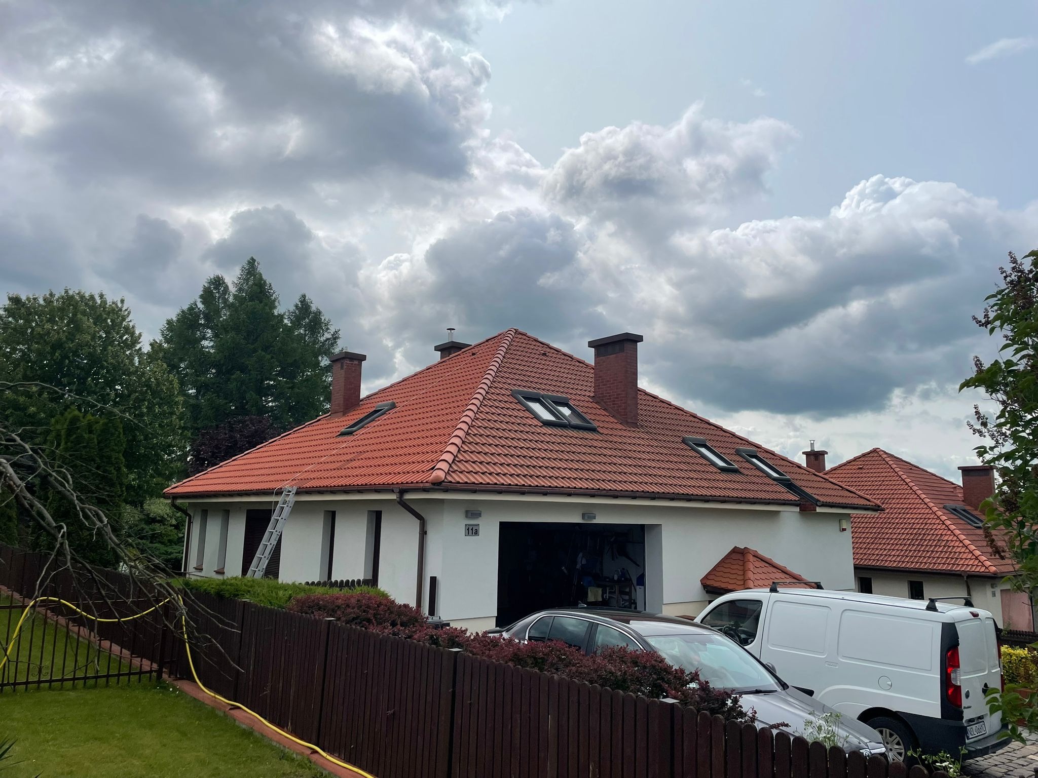 Dom jednorodzinny z czerwoną dachówką, drabina oparta o ścianę, widoczny samochód osobowy i dostawczy. Pochmurne niebo w tle.